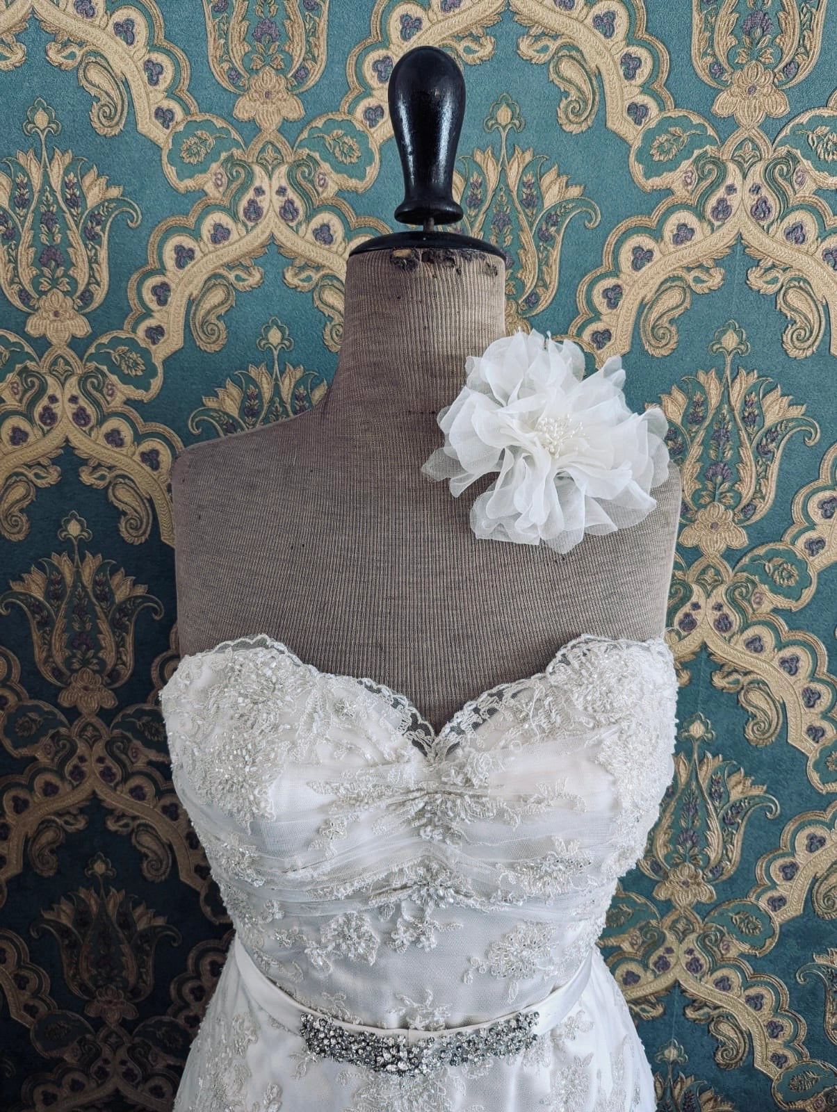 Broochella aubrieta bridal  flower brooch in ivory. Pinned on a mannequin. The mannequin is dressed in a white, beaded, strapless wedding dress. The background is green and gold damask wallpaper.