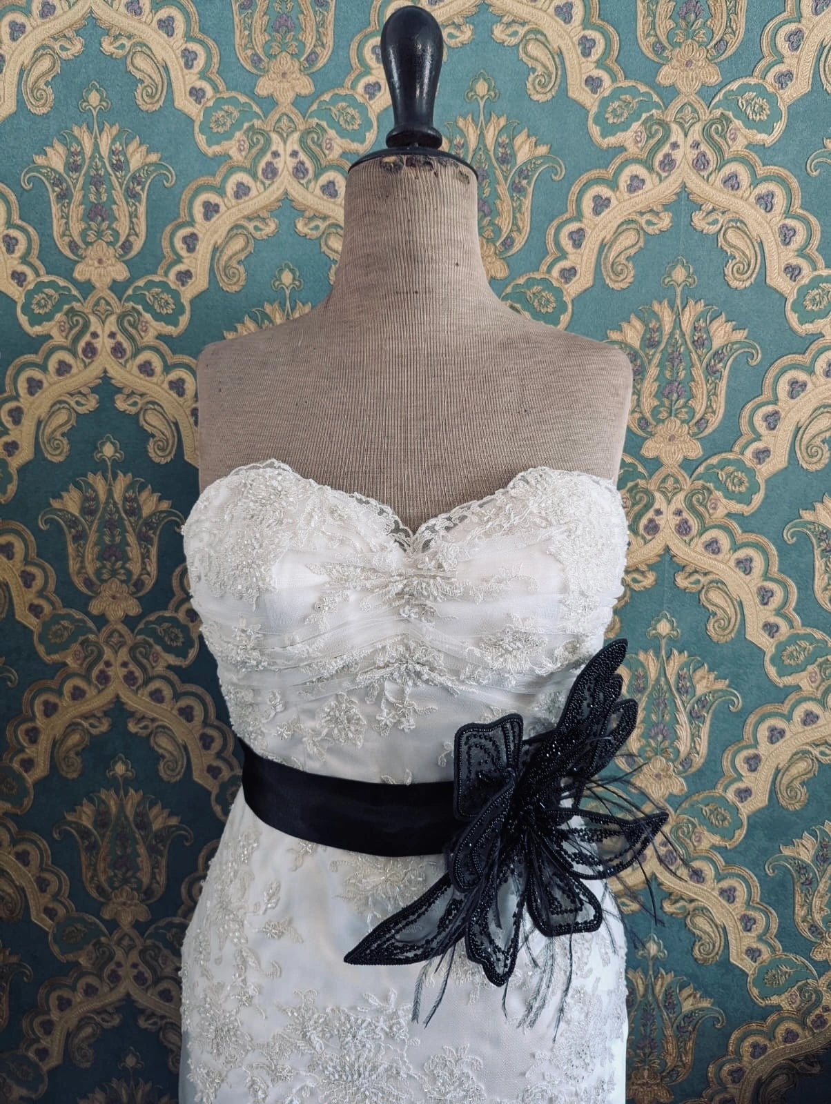 Broochella azalea large bridal, hand beaded, black, large flower brooch, with lace and crystal brooch embellishment. Pinned on a mannequin. The mannequin is dressed in a white, beaded, strapless wedding dress. The background is green and gold damask wallpaper.