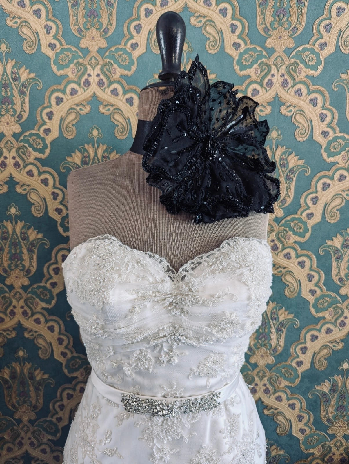 Broochella baccara bridal, hand beaded, black, large flower brooch. Pinned on a mannequin. The mannequin is dressed in a white, beaded, strapless wedding dress. The background is green and gold damask wallpaper.