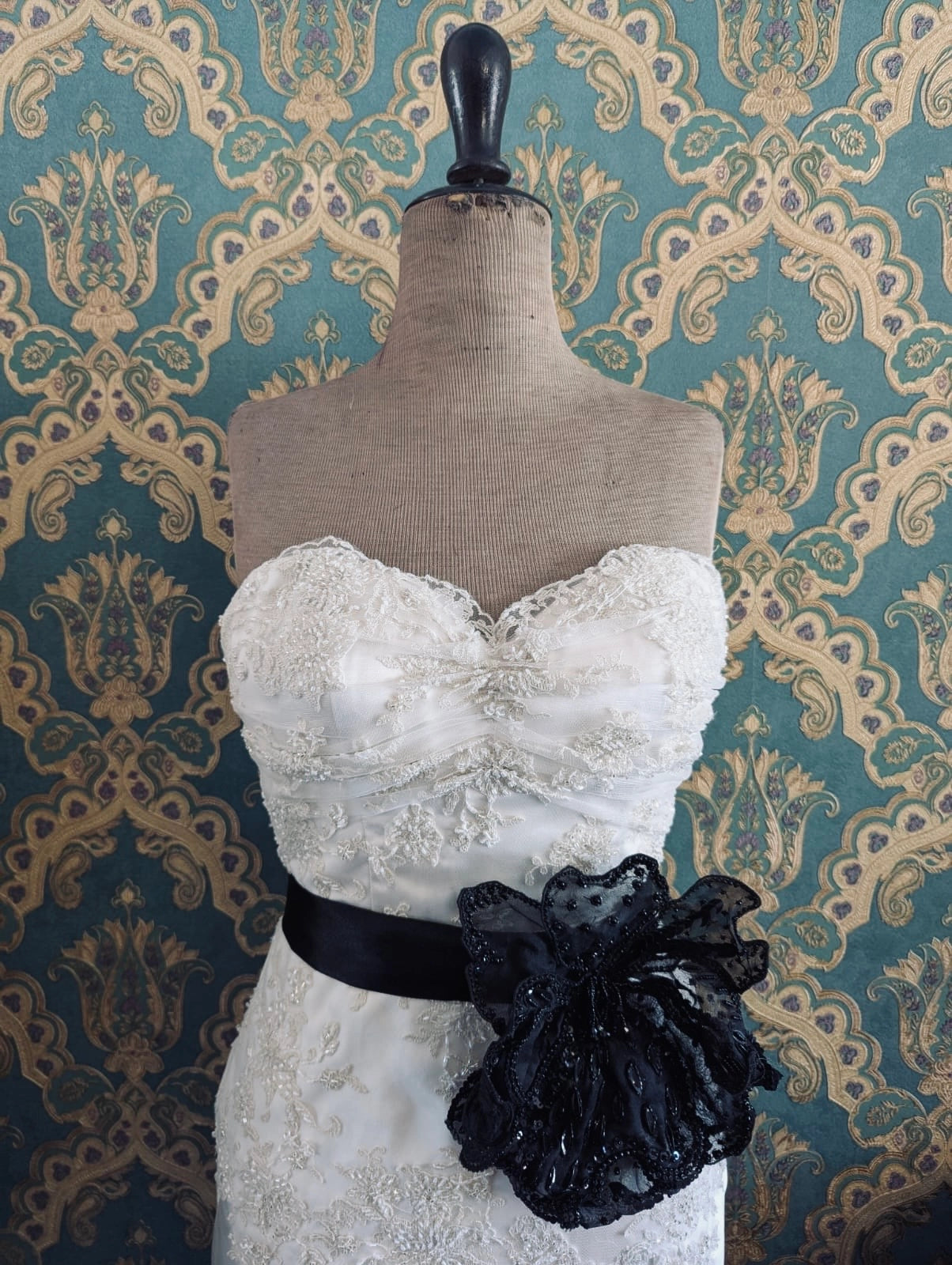Broochella baccara bridal, hand beaded, black, large flower brooch. Pinned on a mannequin. The mannequin is dressed in a white, beaded, strapless wedding dress. The background is green and gold damask wallpaper.