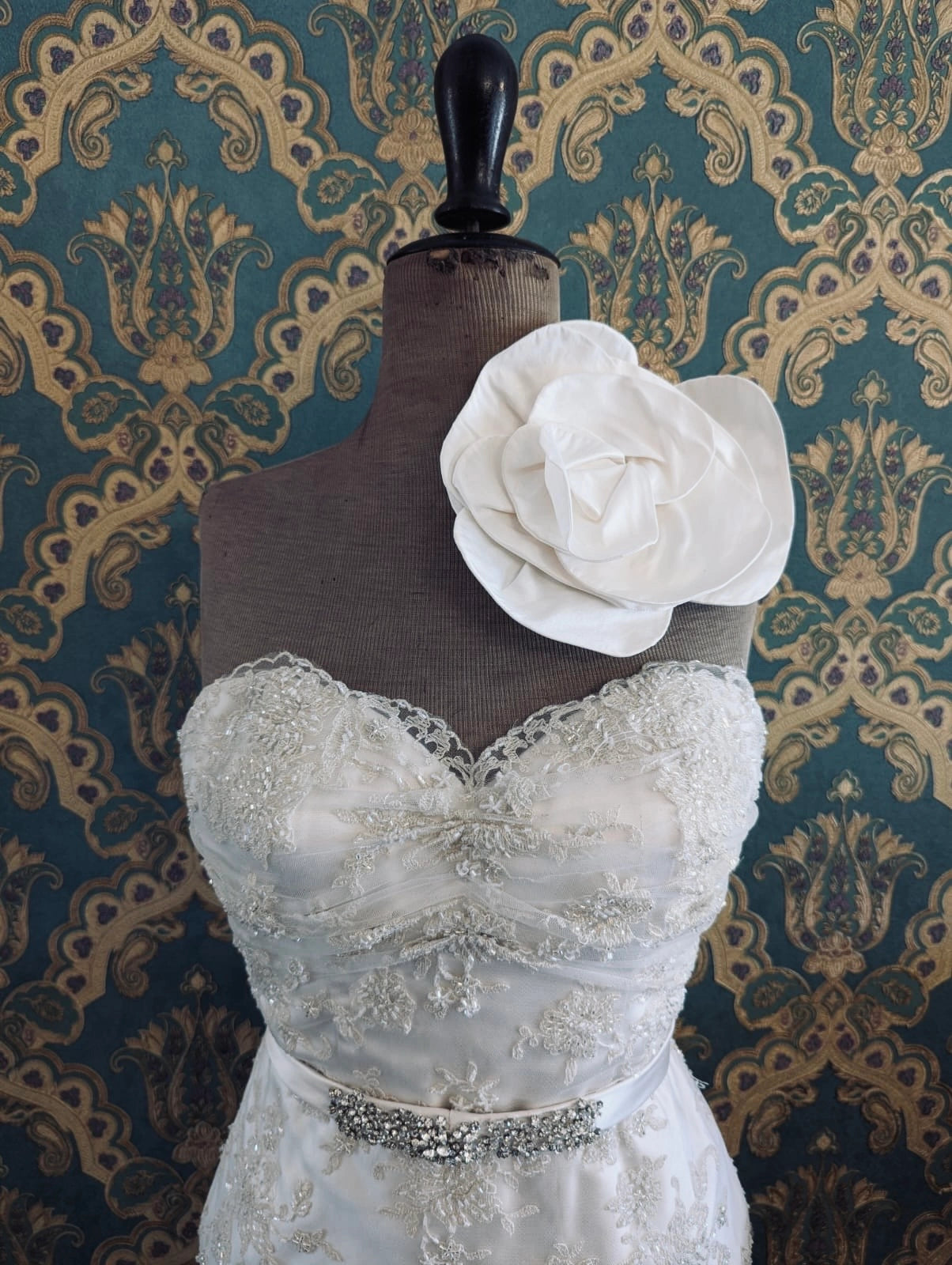 Broochella  bridal handmade bellucci silk flower brooch in ivory. Pictured on a mannequin that is wearing a white, beaded strapless wedding dress. The background is green and gold damask wallpaper. 
