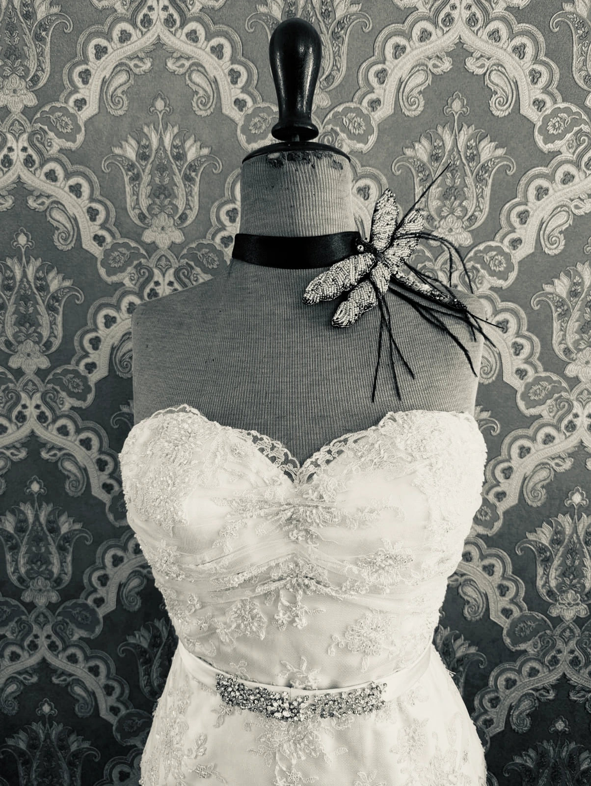 Broochella dragonfly hand beaded pink and black bridal brooch. Pictured on a mannequin that is wearing a white, beaded strapless wedding dress. The background is green and gold damask wallpaper. 