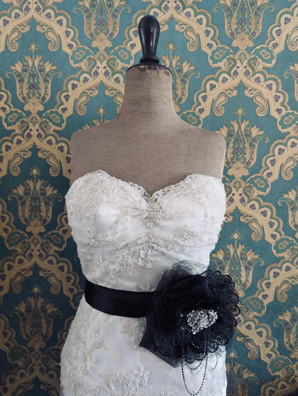 Broochella madonna bridal, hand beaded, black, large flower brooch, with lace and crystal brooch embellishment. Pinned on a mannequin. The mannequin is dressed in a white, beaded, strapless wedding dress. The background is green and gold damask wallpaper.