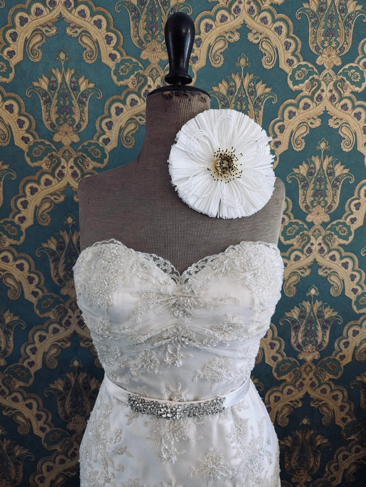 Broochella bridal raffia handmade flower brooch with embellished centre . Pictured on a mannequin that is wearing a white, beaded strapless wedding dress. The background is green and gold damask wallpaper.