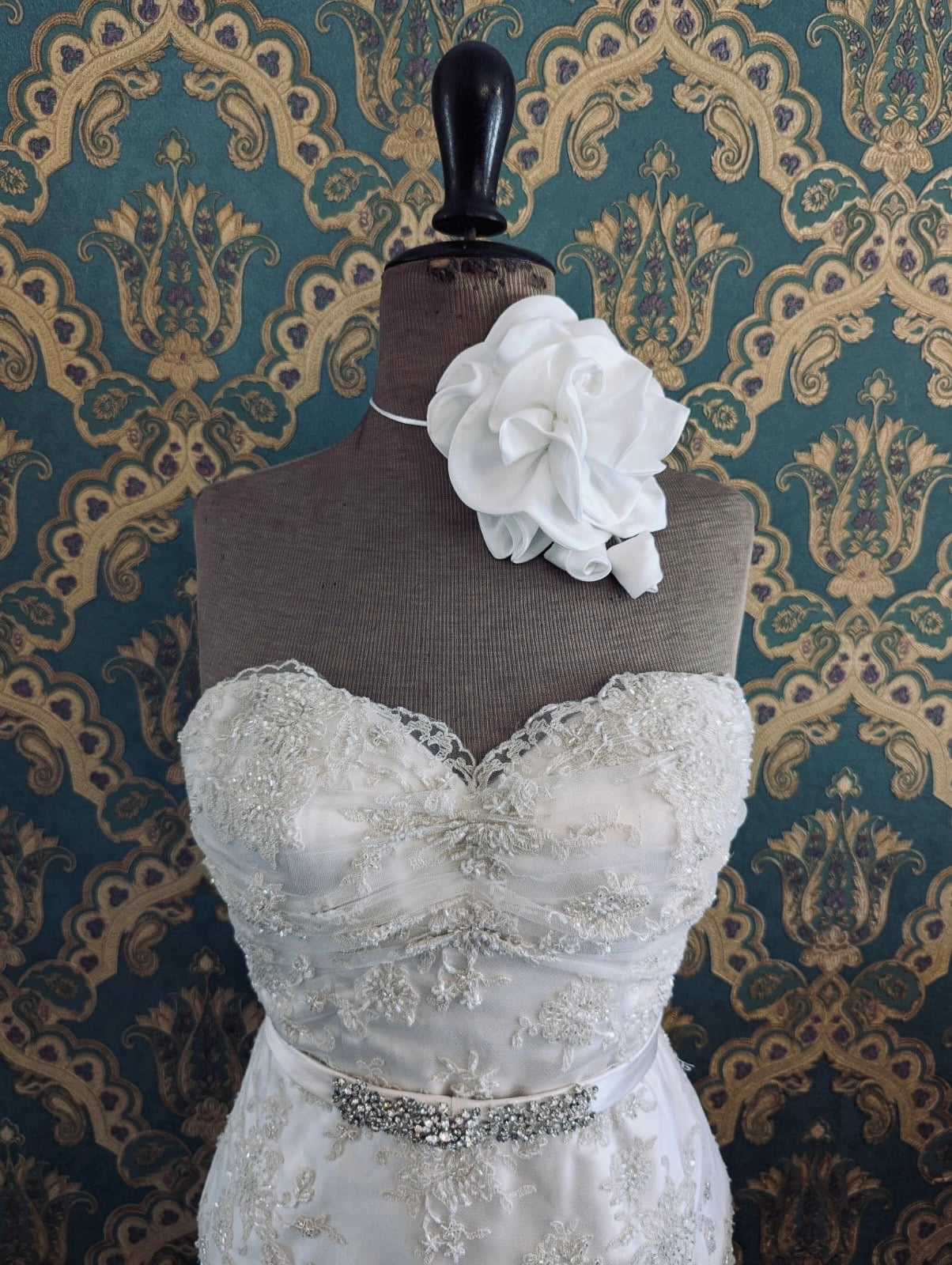 Broochella rosetta white handmade designer bridal flower choker and brooch. Pictured on a mannequin that is wearing a white, beaded strapless wedding dress. The background is green and gold damask wallpaper. 