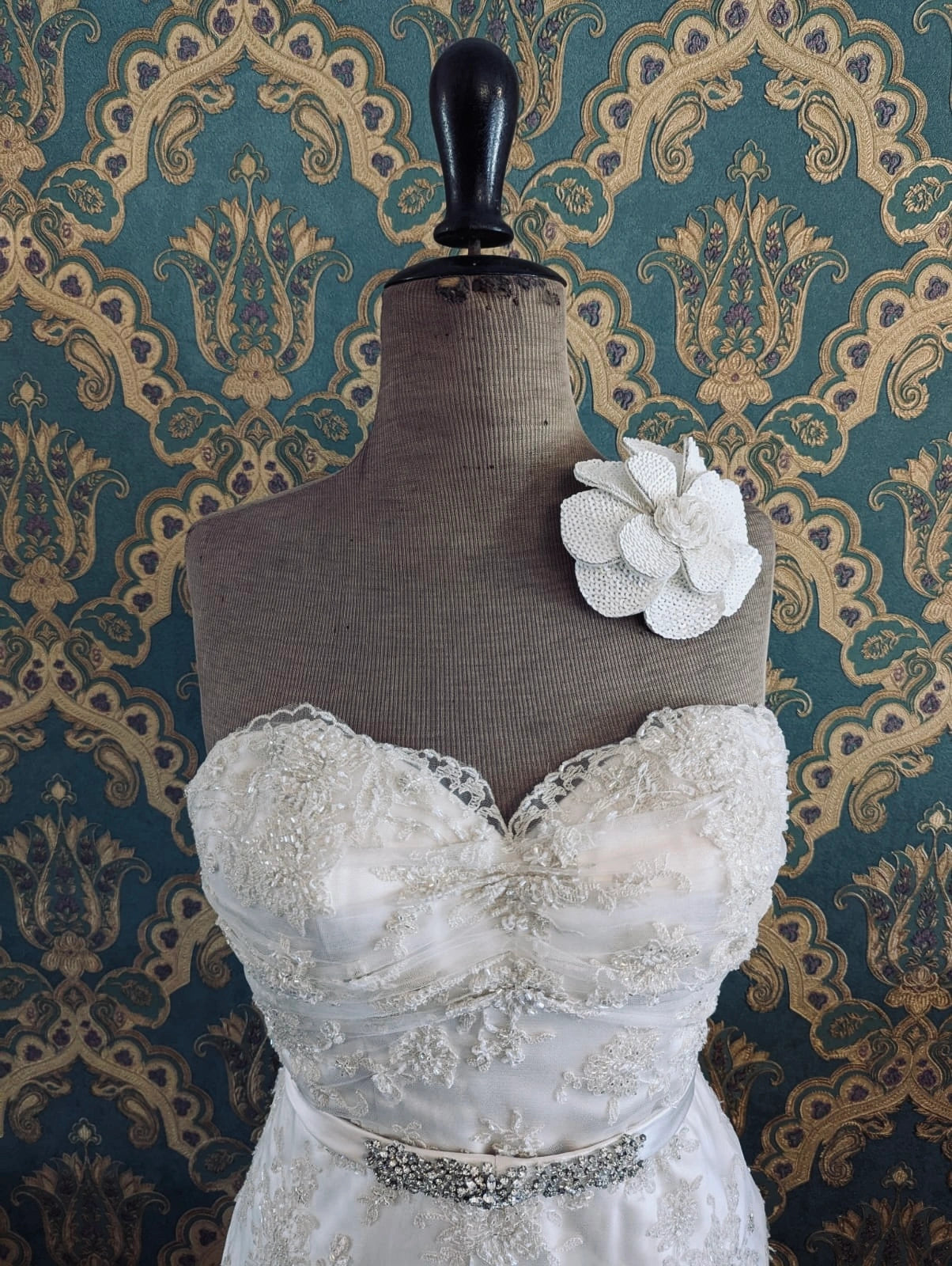 Broochella bridal scintillante hand beaded, white sequin wedding flower brooch. Pictured on a mannequin that is wearing a white, beaded strapless wedding dress. The background is green and gold damask wallpaper. 