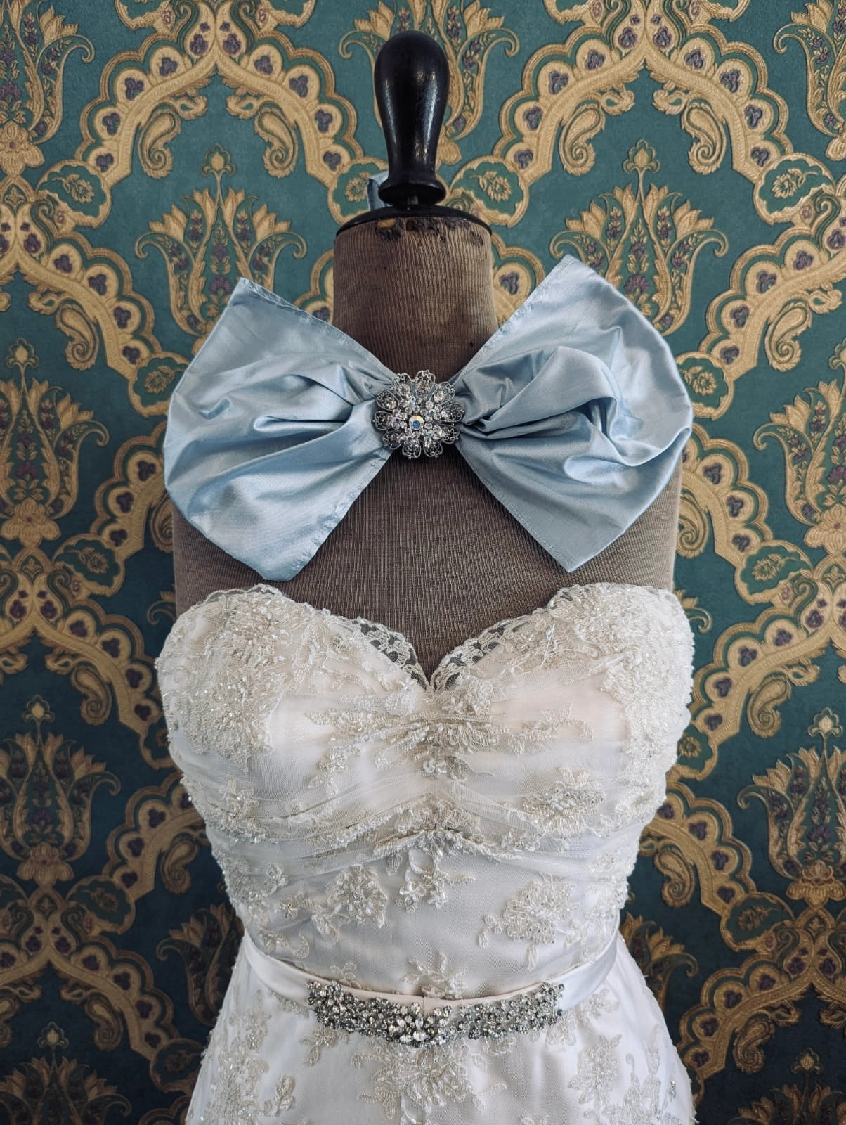 Broochella bridal silk something blue handmade bow brooch with a silver crystal brooch embellishment at the centre. Pictured on a mannequin that is wearing a white, beaded strapless wedding dress. The background is green and gold damask wallpaper. 