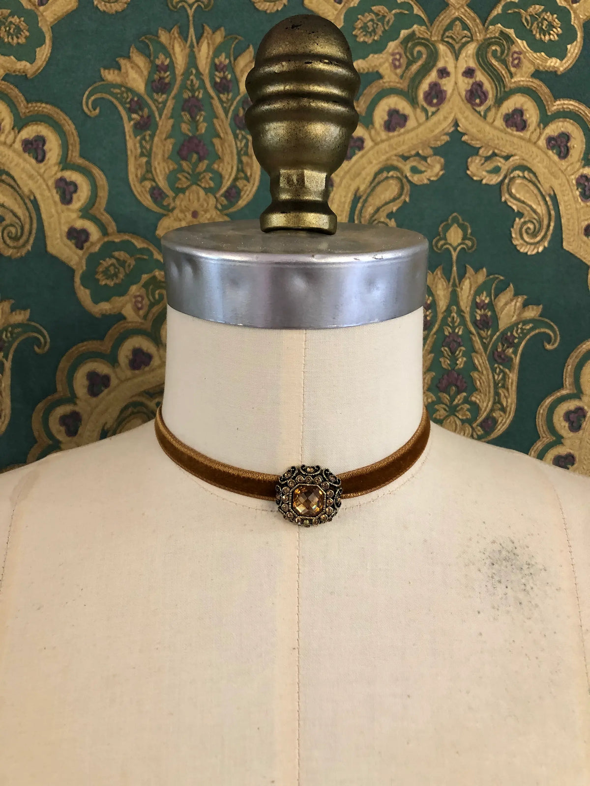 A Broochella Lady Whistledown bronze velvet choker necklace with a decorative antique bronze filigree centrepiece, displayed on a cream-coloured dress form against a patterned teal and gold damask wallpaper background.