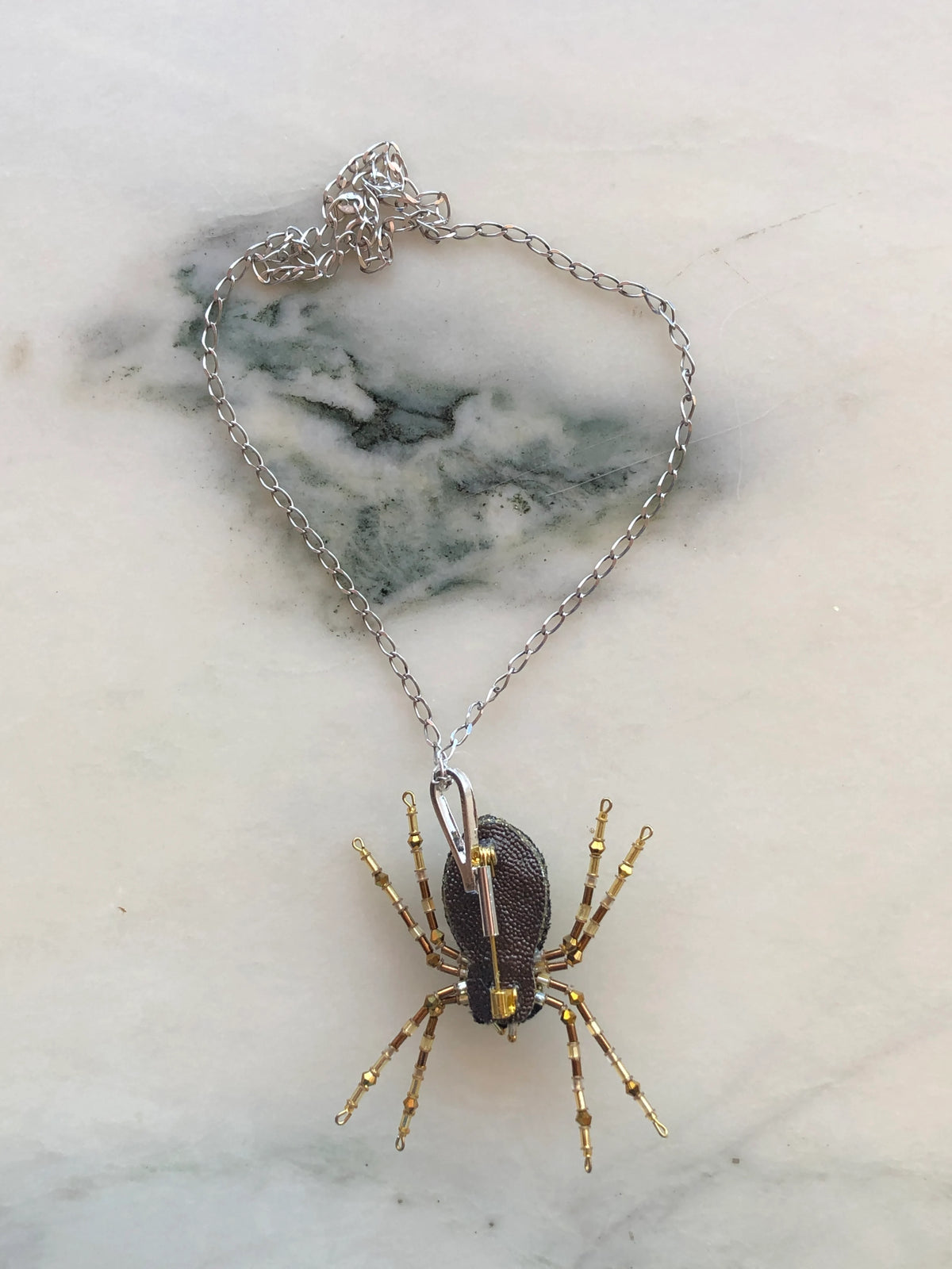 Broochella vertical brooch to necklace converter in silver-tone metal with elongated loop design. It is pictured in use on the Broochella spider brooch and a silver chain. It is photographed on white marble with grey-green veining.