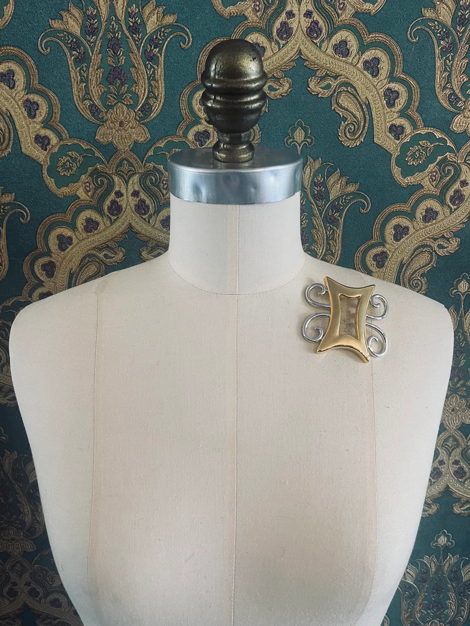 Broochella brooches with meaning strong and humble gold and silver metal brooch. Pictured on a mannequin. The background is green and gold damask wallpaper