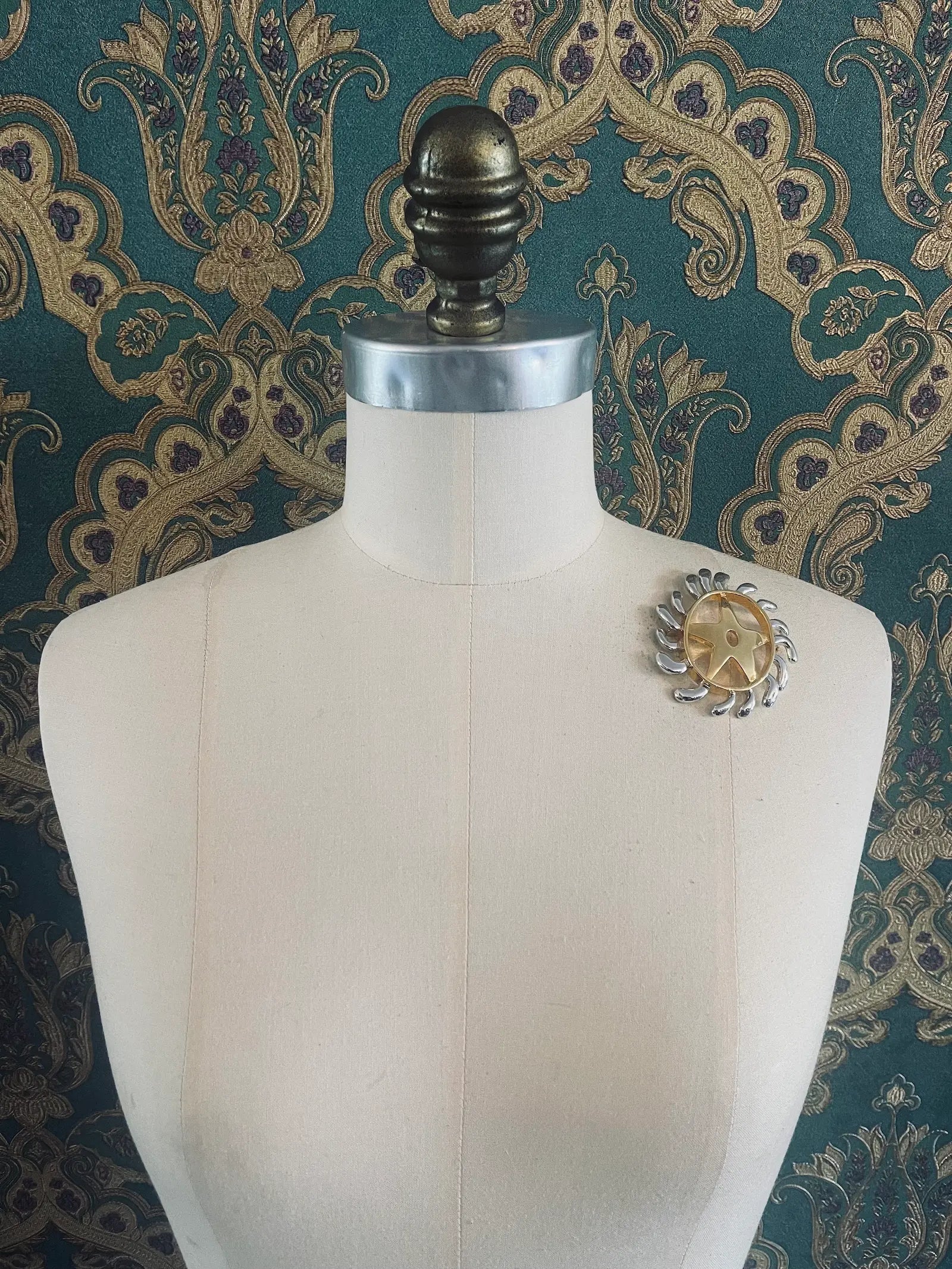 Broochella brooches with meaning welcoming change gold and silver metal brooch. Pictured on a mannequin. The background is green and gold damask wallpaper