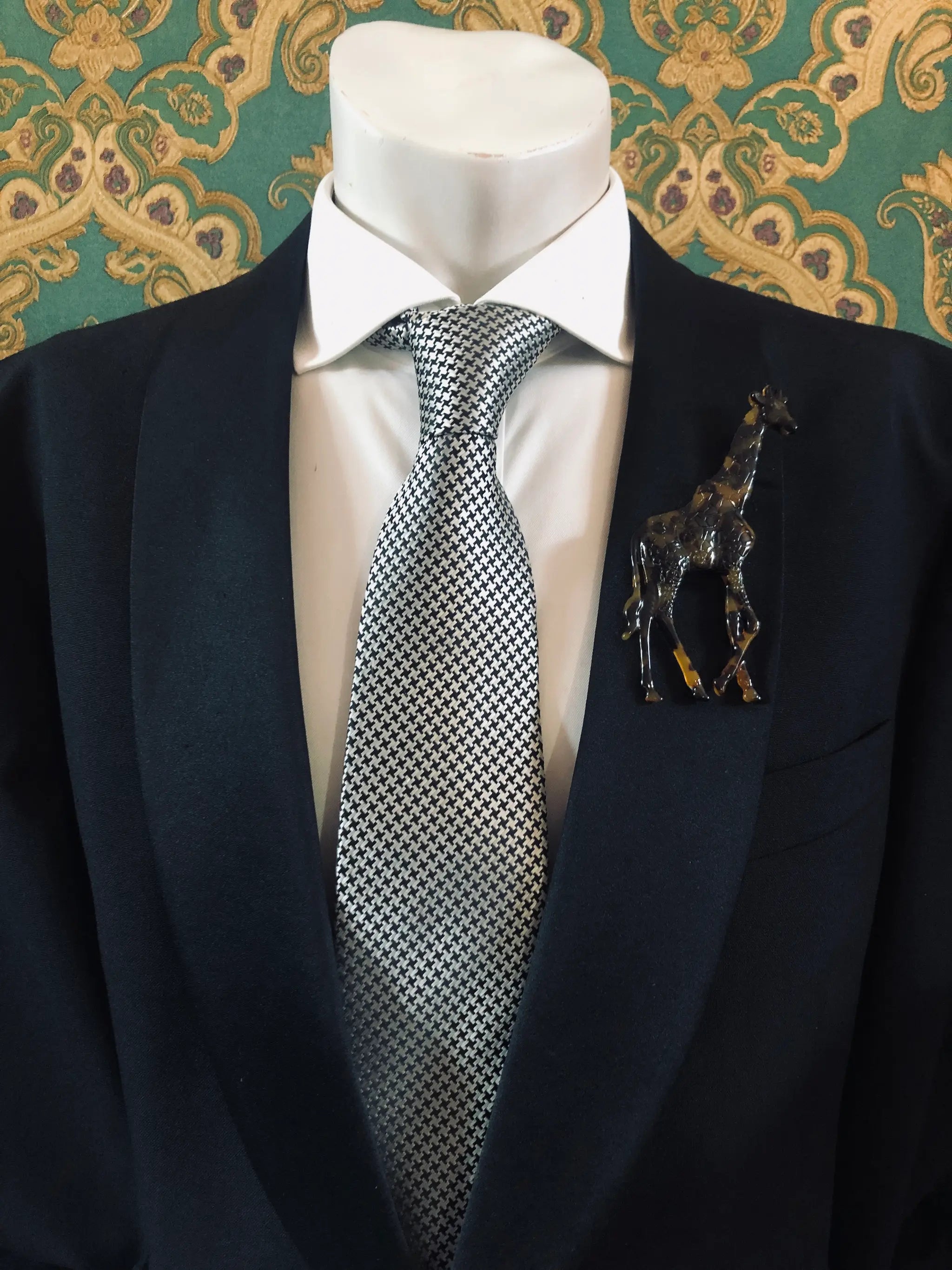 Broochella Men's brown Animalia tortoiseshell giraffe shaped brooch. It is made from Premium Italian cellulose tortoiseshell acetate. It is presented on a men's mannequin that is dressed in a black suit. The background is green and gold damask wallpaper. 