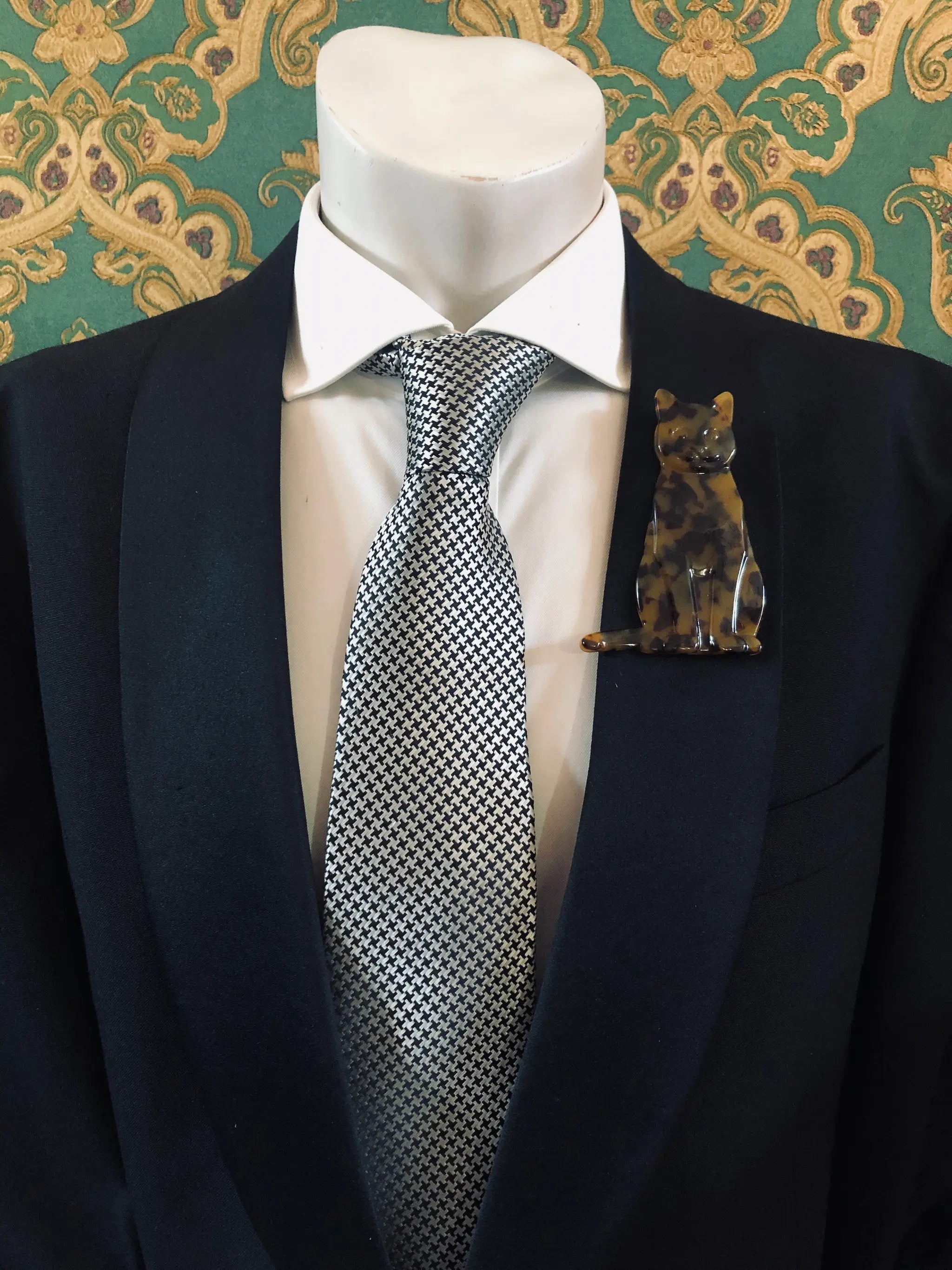 Broochella Men's brown Animalia tortoiseshell giraffe shaped brooch. It is made from Premium Italian cellulose tortoiseshell acetate. It is presented on a men's mannequin that is dressed in a black suit. The background is green and gold damask wallpaper. 