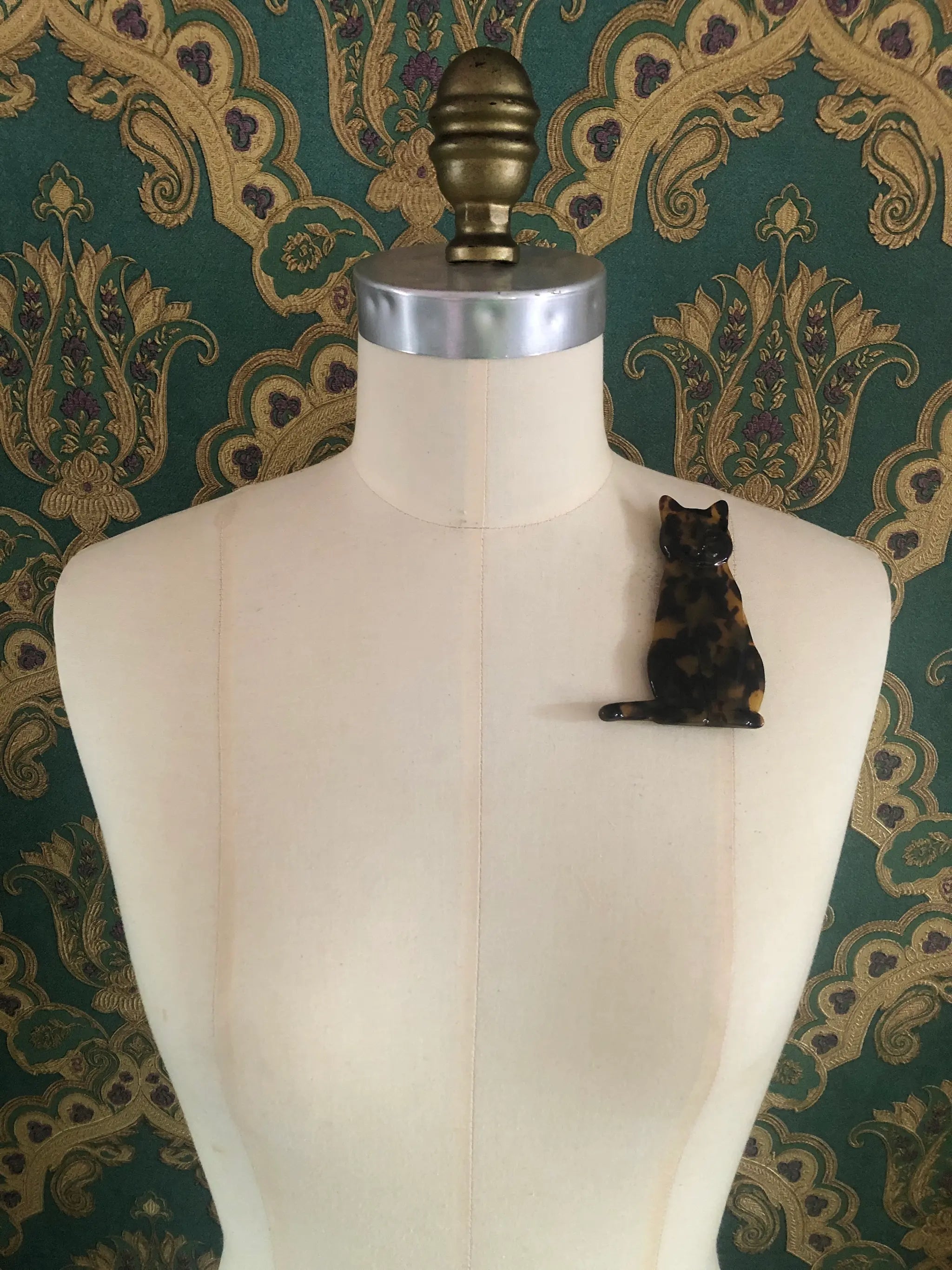 A Broochella Animalia brown tortoiseshell cat-shaped brooch made from premium Italian cellulose acetate. It  displayed on a mannequin. The background is green and gold damask wallpaper. 