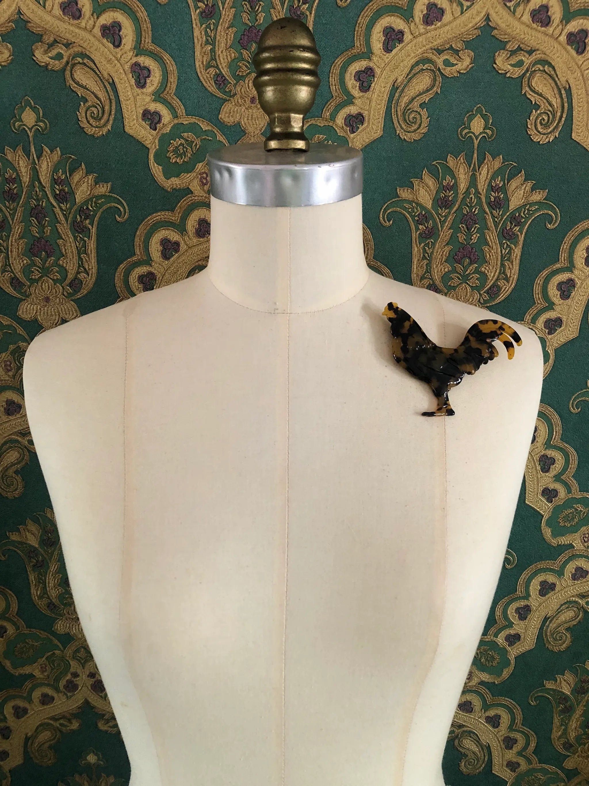 Broochella Animalia handmade, tortoiseshell, Italian cellulose acetate rooster brooch in brown. It is pinned to a mannequin. The background is green and gold damask wallpaper.