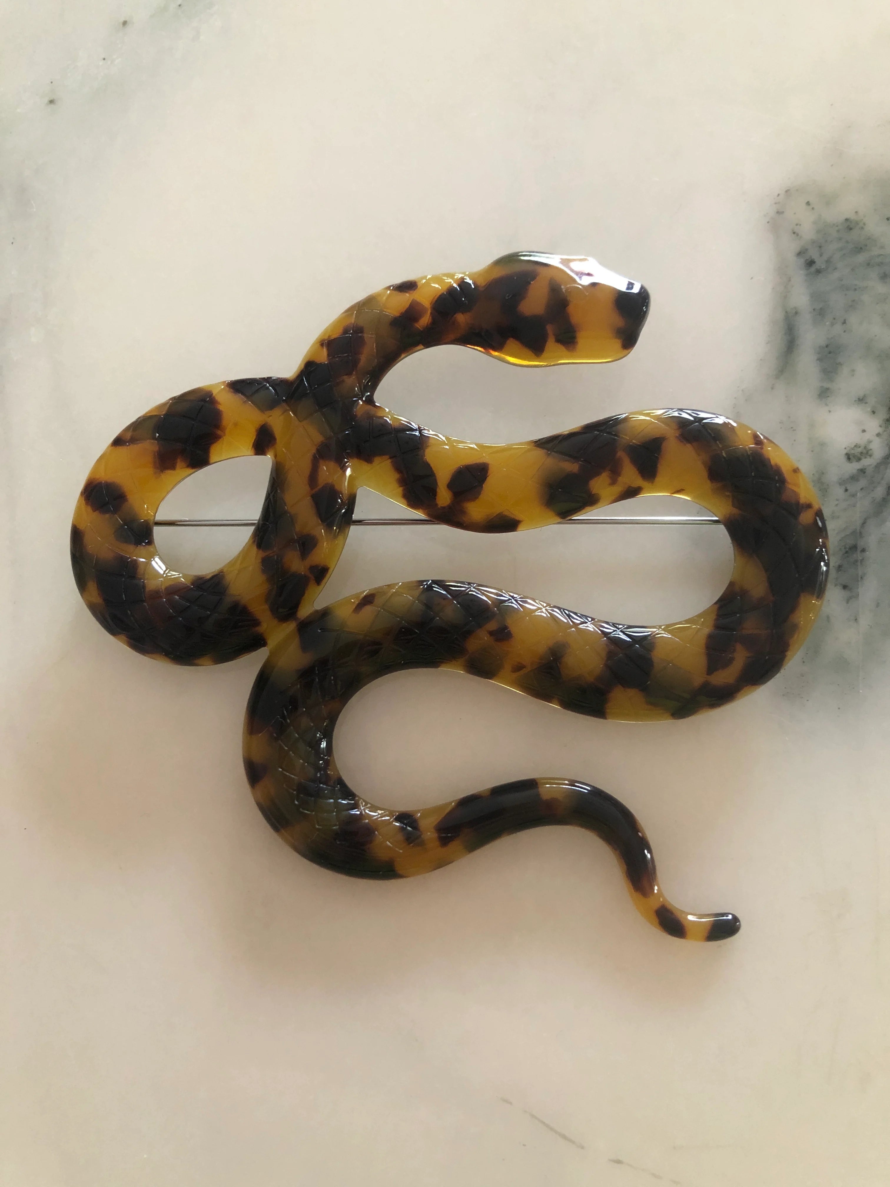 A Broochella Animalia brown tortoiseshell snake-shaped brooch made from premium Italian cellulose acetate. It  displayed on a white marble table