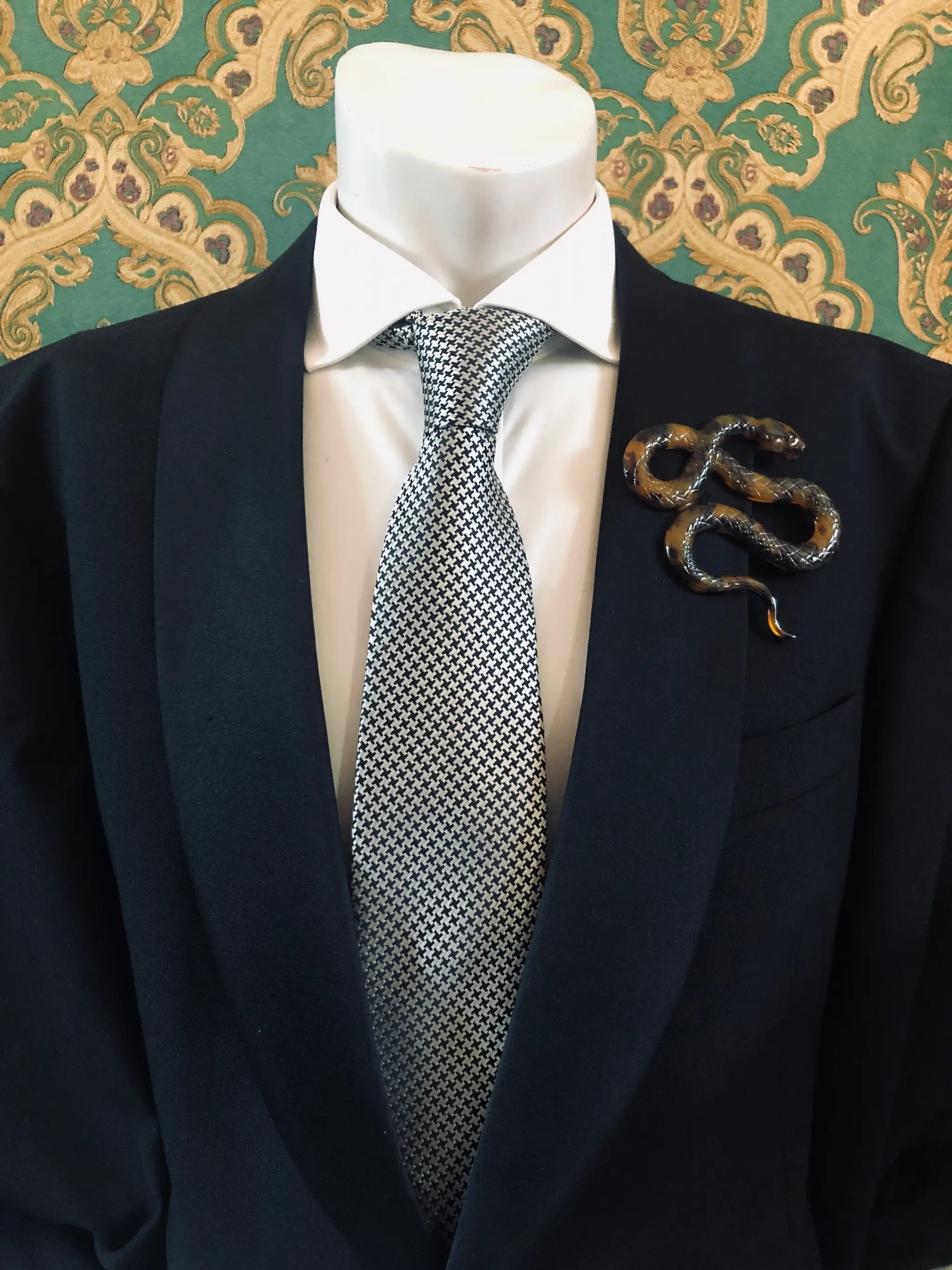 Broochella Men's brown Animalia tortoiseshell giraffe shaped brooch. It is made from Premium Italian cellulose tortoiseshell acetate. It is presented on a men's mannequin that is dressed in a black suit. The background is green and gold damask wallpaper. 