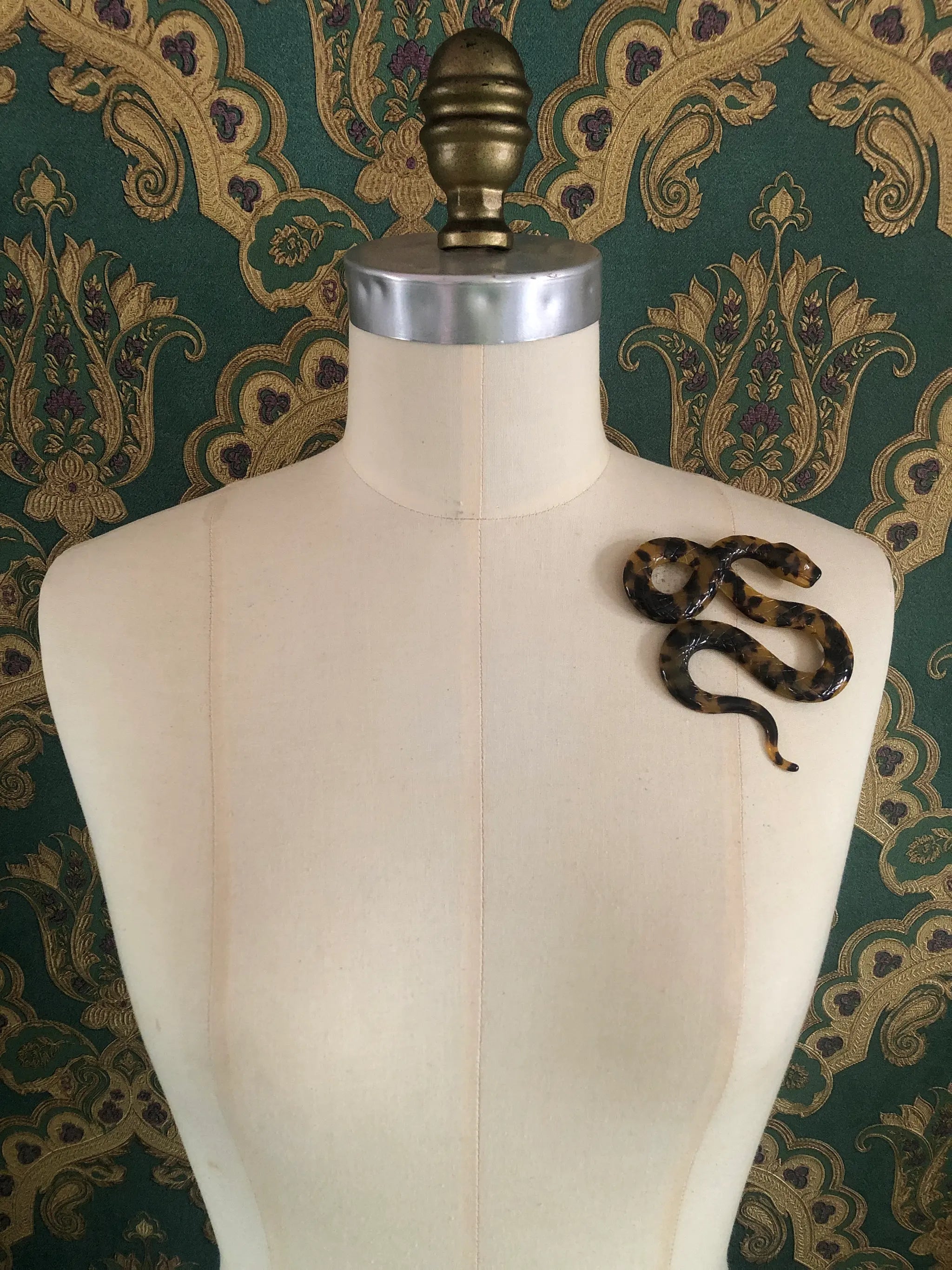 A Broochella Animalia brown tortoiseshell snake-shaped brooch made from premium Italian cellulose acetate. It  displayed on a cream fabric dress form against a green and gold patterned wallpaper background.