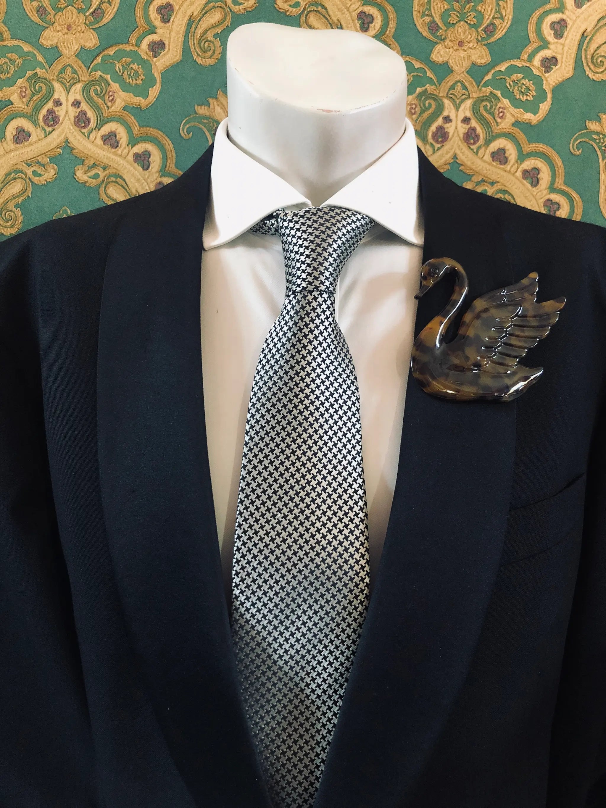 A Broochella Animalia Men's brown tortoiseshell rooster brooch displayed on a white mannequin torso, featuring a black  suit jacket, a white dress shirt, a silver and black checkered tie against a green and gold patterned wallpaper background.