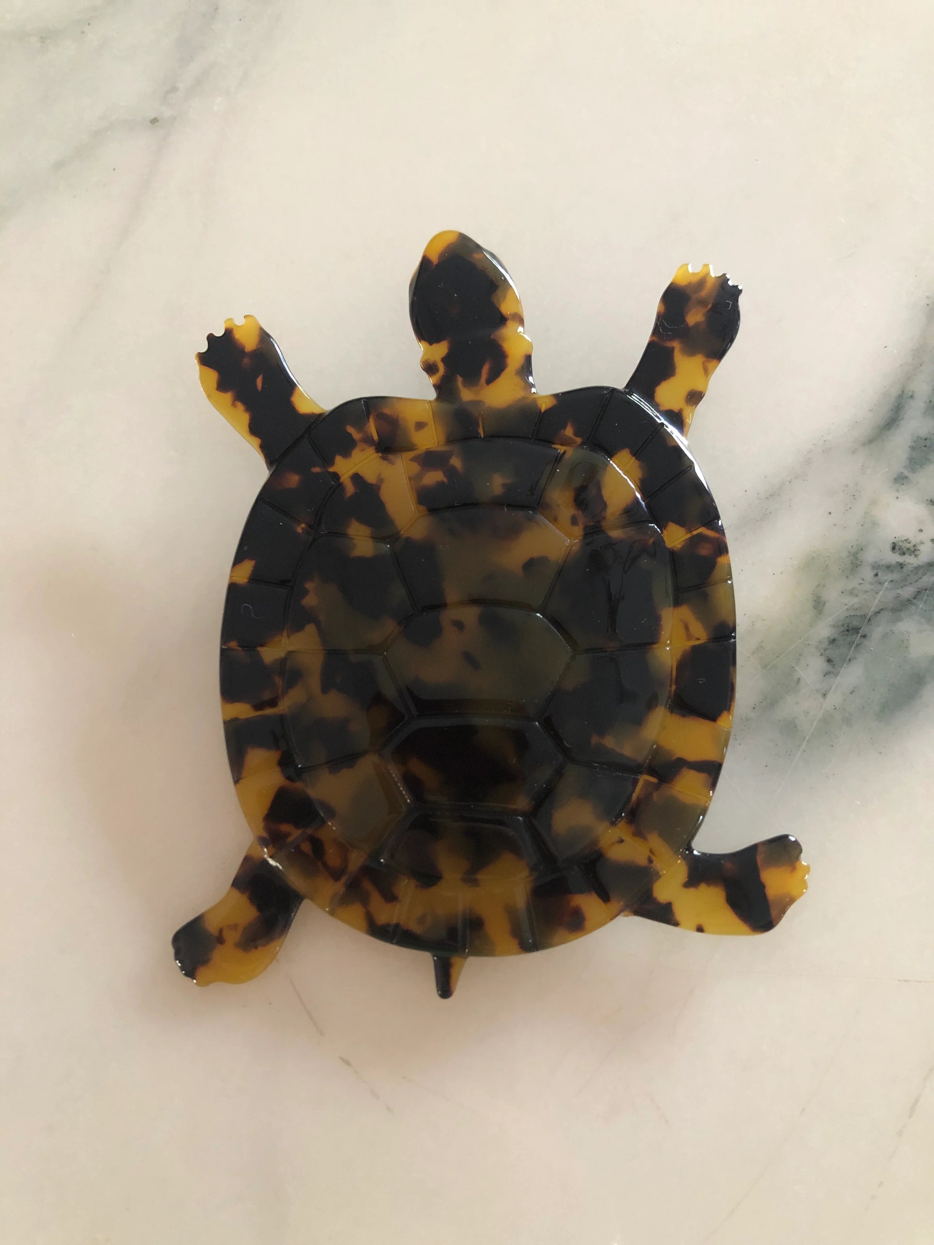 A Broochella Animalia Collection tortoise brooch in black, beige and brown tortoiseshell Italian cellulose acetate with detailed shell plate texture measuring 10cm x 8cm. The background is white marble. 