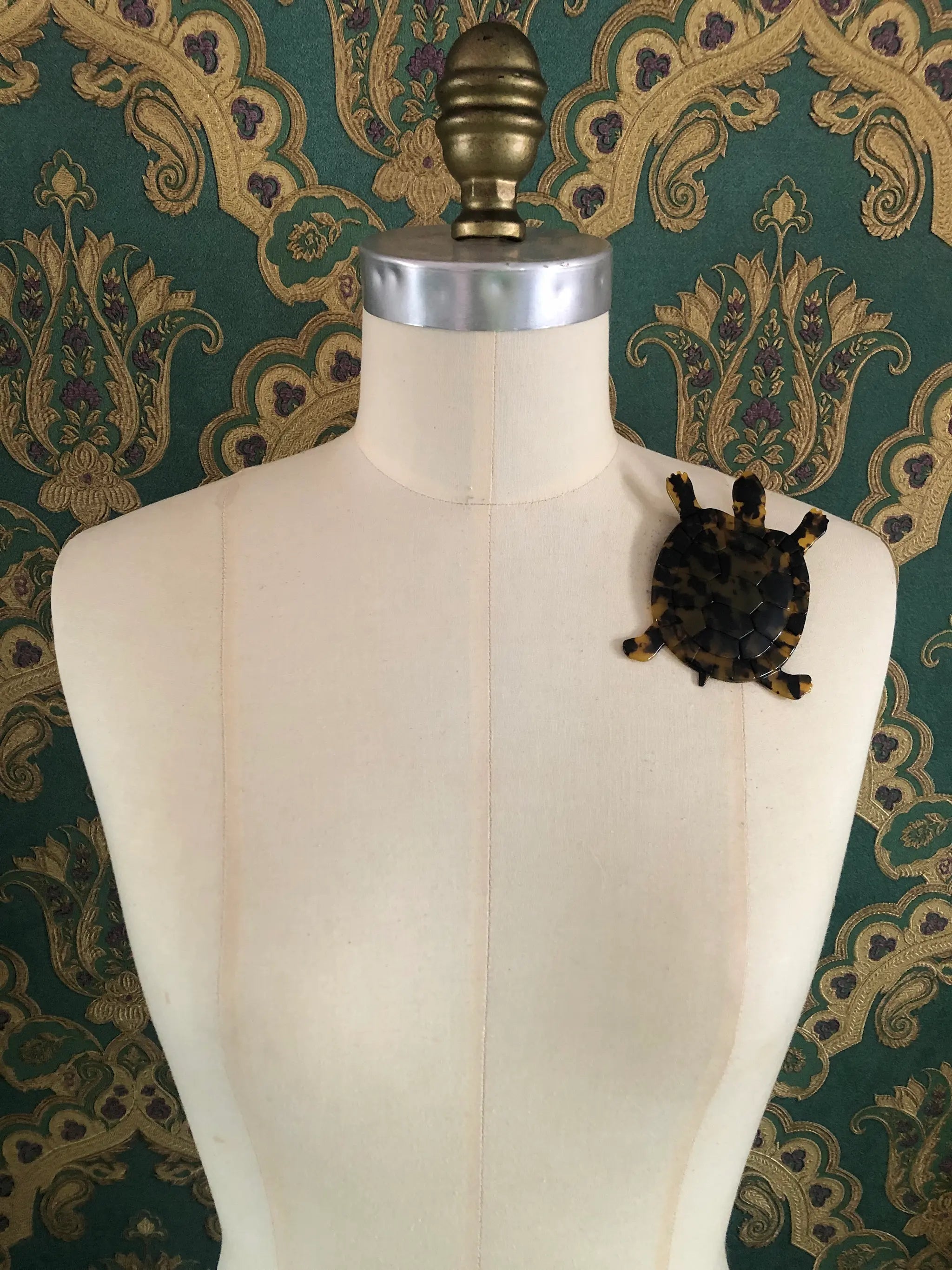 A Broochella Animalia Collection tortoise brooch in black and brown tortoiseshell Italian cellulose acetate with detailed shell plate texture measuring 10cm x 8cm. It is pinned to a mannequin. The background is green and gold damask wallpaper. 