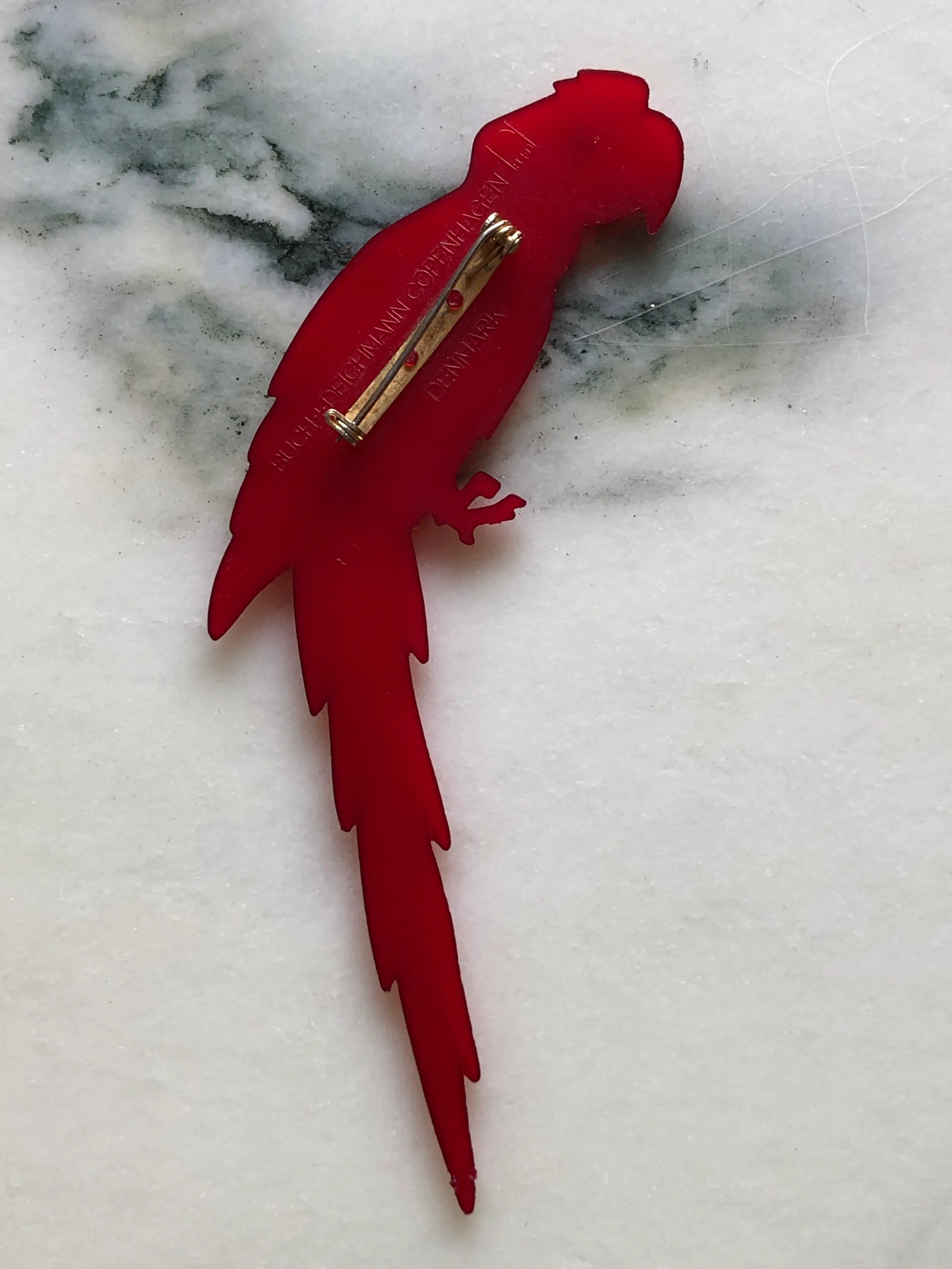 Broochella vintage Buch + Deichmann Scarlet Macaw brooch in vivid red with a clear crystal eye and long sculptural tail, made in Copenhagen, Denmark and measuring approximately 14 × 6 cm. Displayed on green velvet in an ornate gold-tone frame.