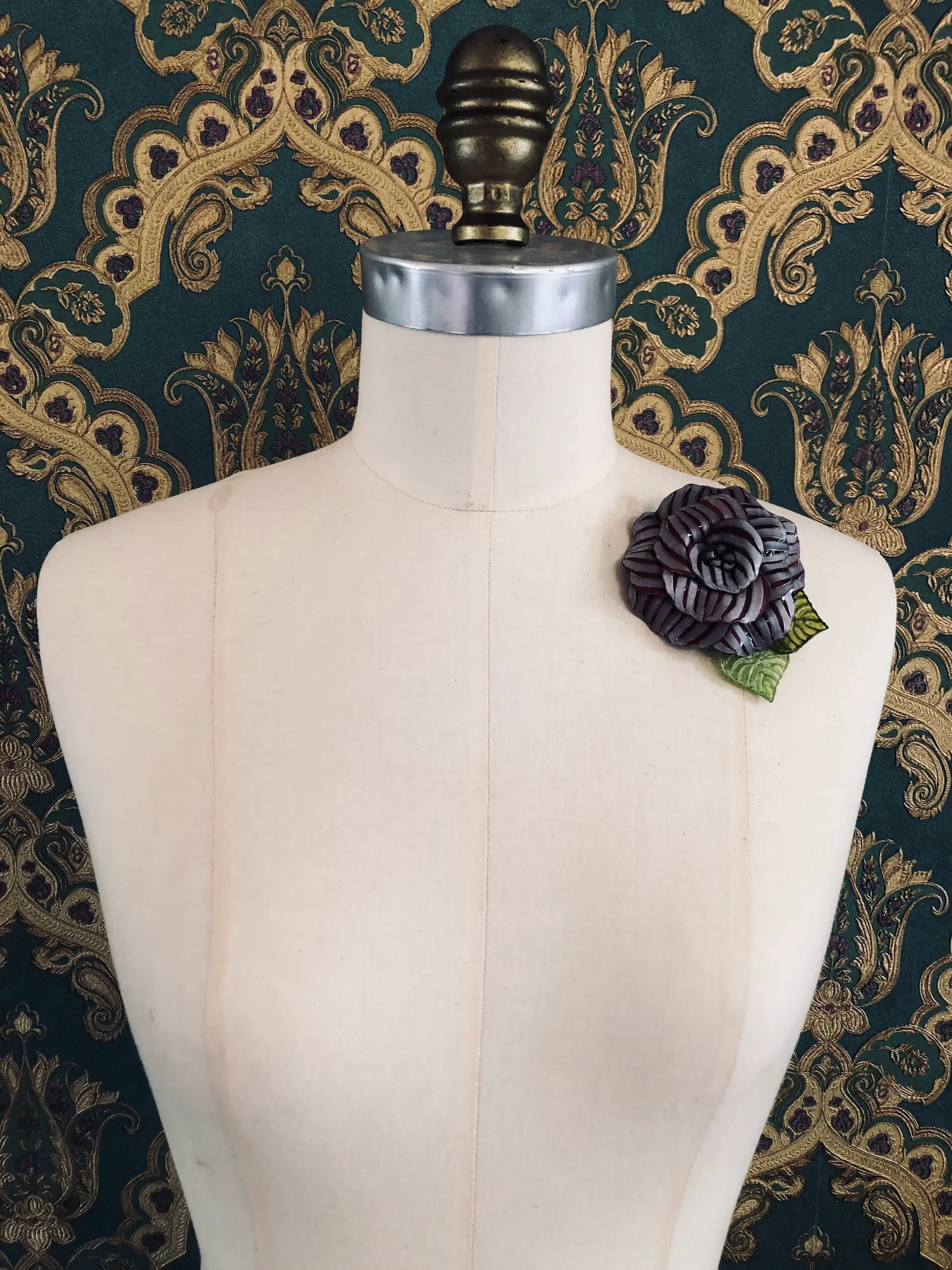A Broochella x Fabrice Paris burgundy moulded plastic rose brooch with green leaves, pinned to the chest of a cream-coloured dress form against a dark green and gold patterned damask backdrop.
