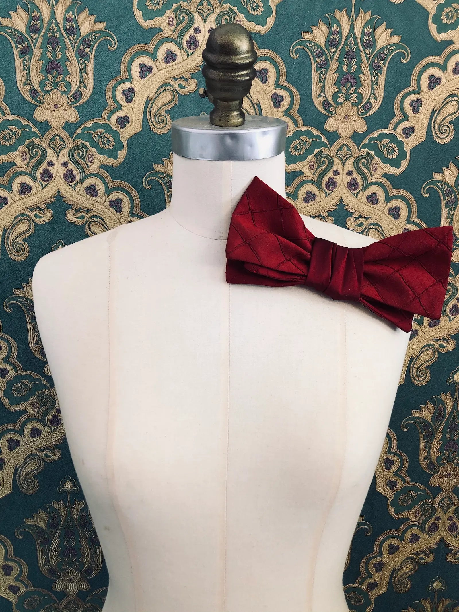 Broochella Marie Antoinette handmade, designer silk bow brooch in burgundy. It is pinned to a mannequin. The background is green and gold damask wallpaper.