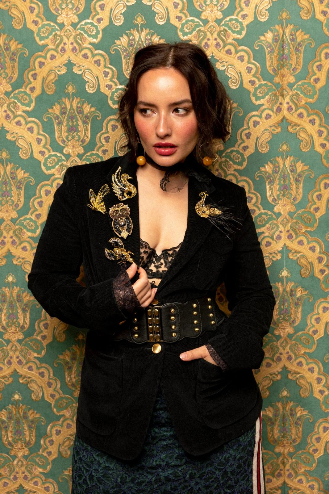 A woman posing in front of ornate green and gold damask wallpaper, wearing a black velvet blazer with gold embroidery, a wide black belt with gold studs, and a patterned skirt. She is wearing The Broochella butterfly brooch 