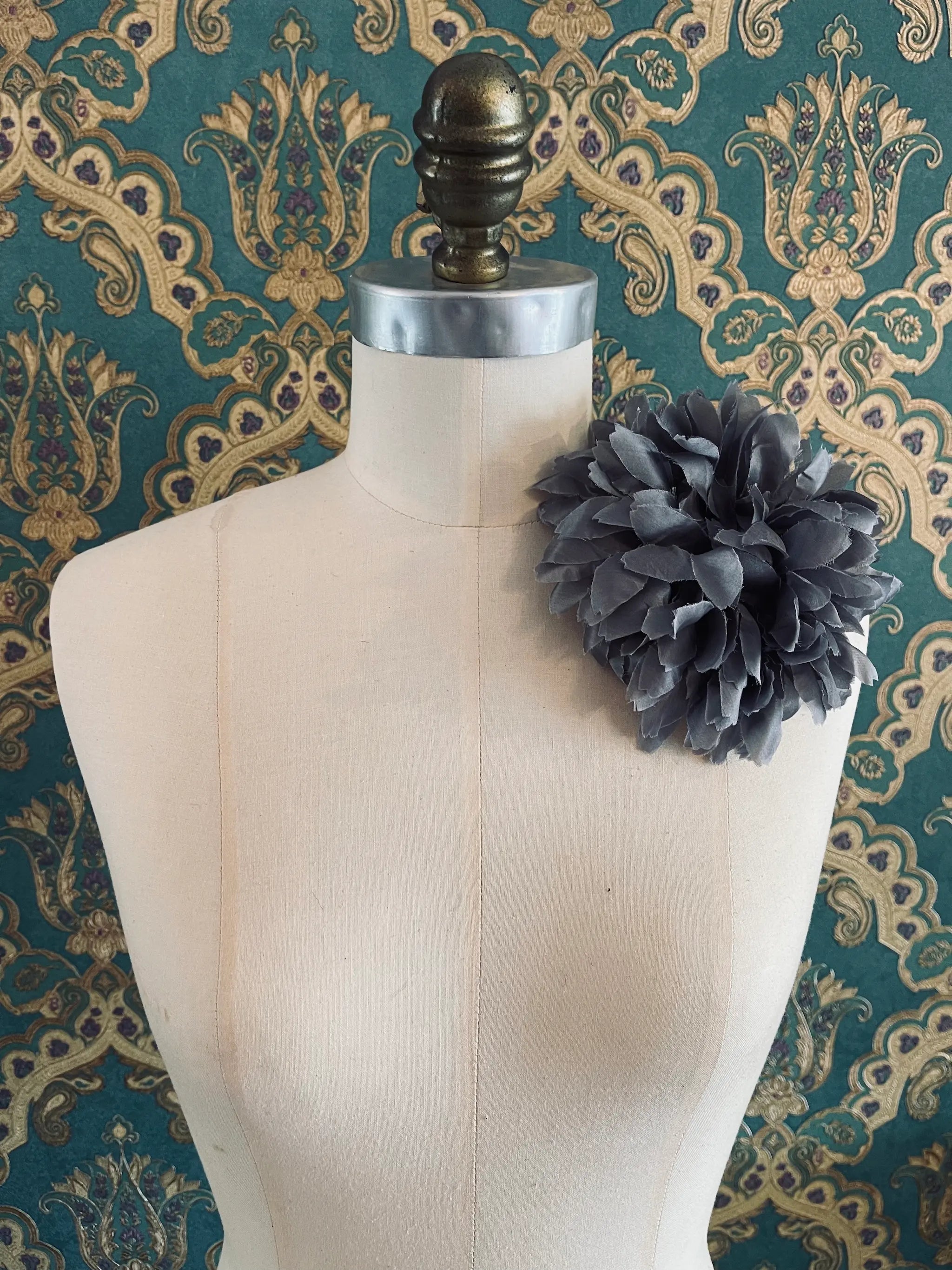 Broochella Calendula handmade, grey fabric flower brooch. Styled on a mannequin. The background is green and gold damask wallpaper