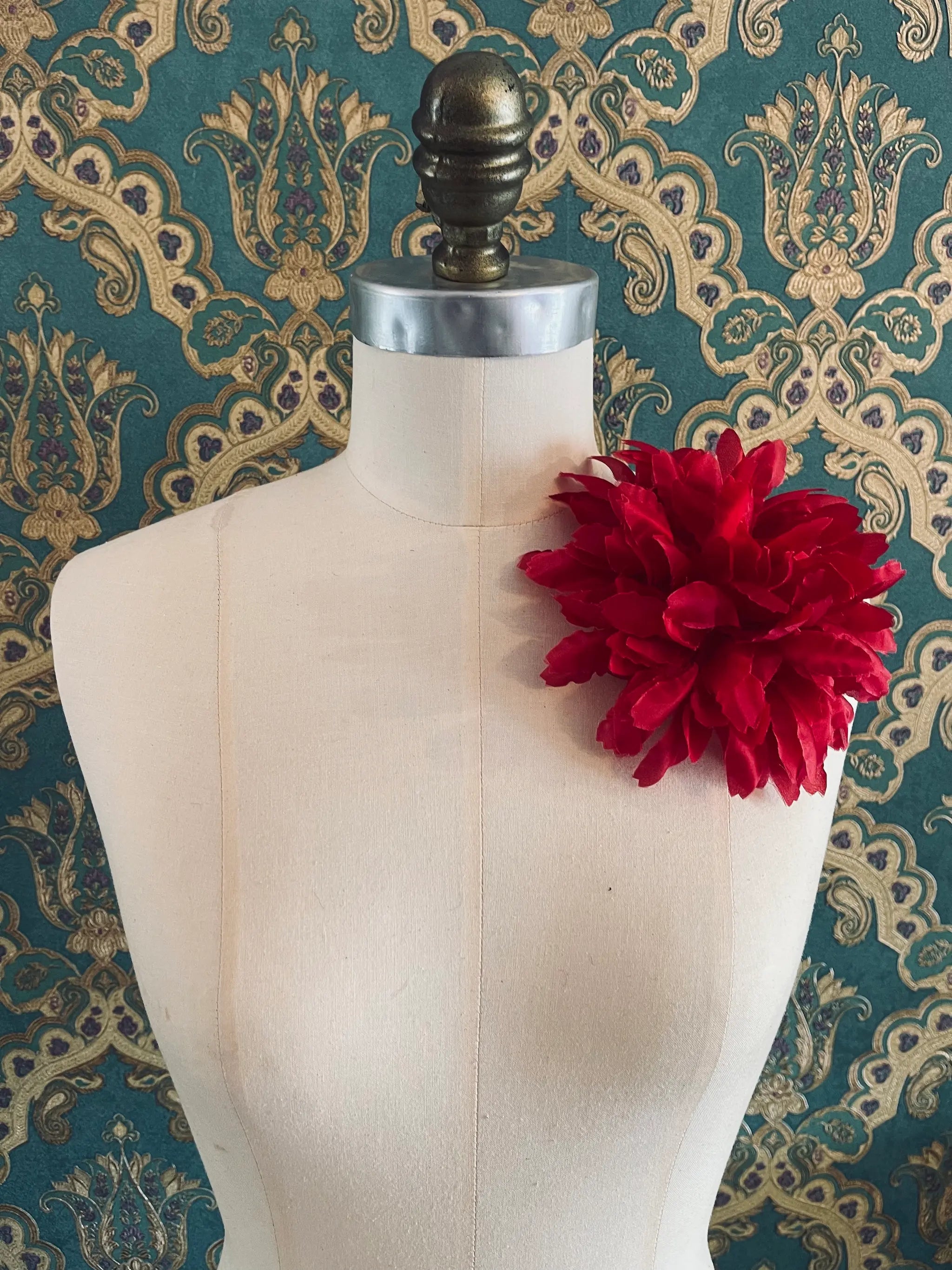 Broochella calendula red handmade fabric designer flower brooch. Styled on a mannequin. The background is green and gold damask wallpaper