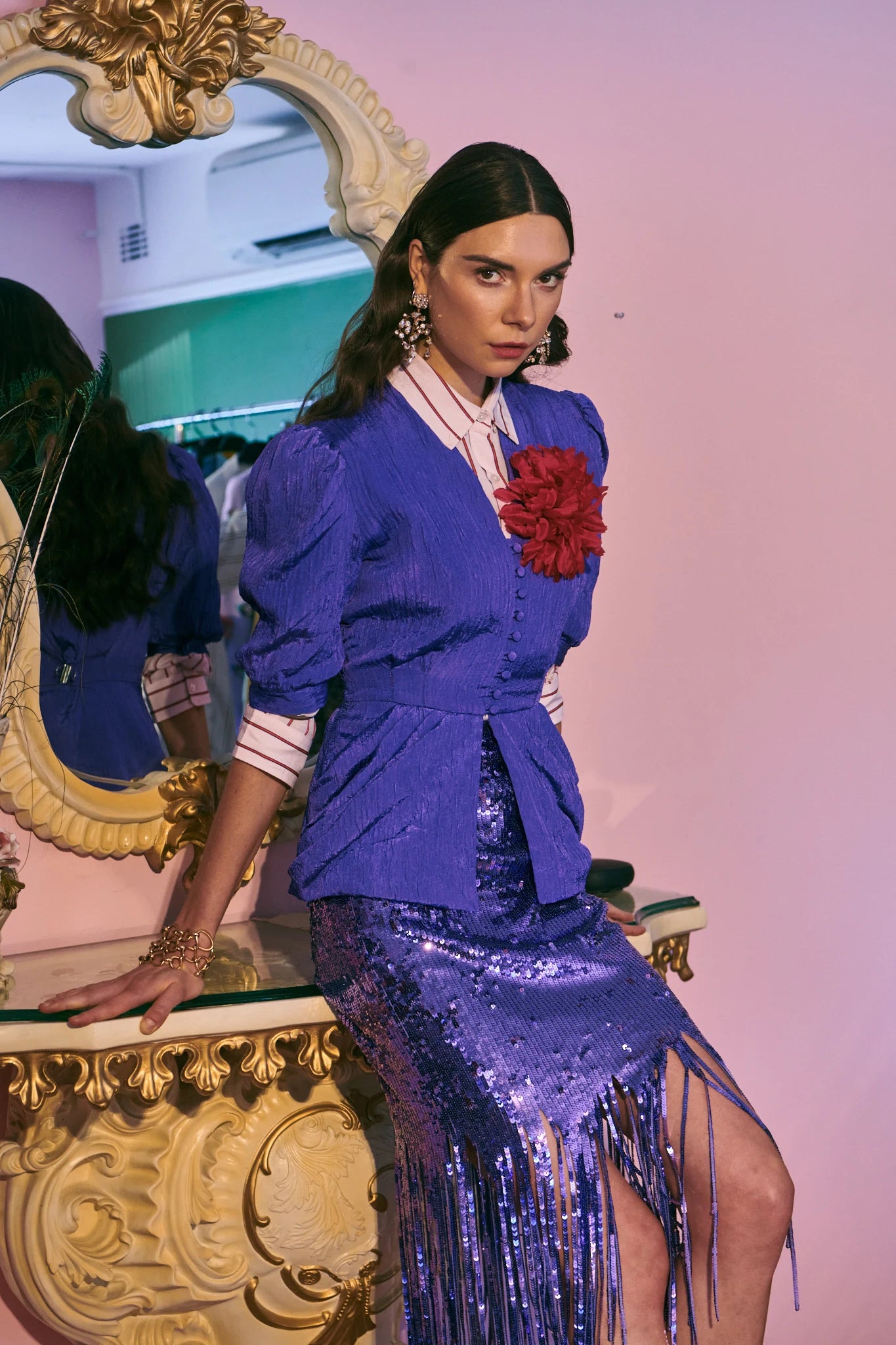 A model wearing a vibrant purple two-piece outfit consisting of a puffed jacket with a Broochella red calendula flower brooch and a matching fringe skirt, posed in front of a pink wall and an ornate gold mirror.