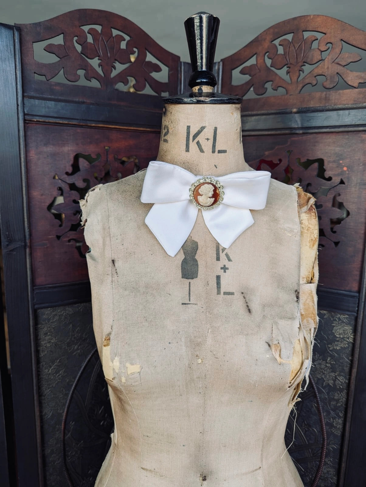 Broochella camee ivoire ivory handmade satin bow brooch with vintage cameo brooch embellishment. It is pinned to a vintage mannequin. The background is a decorative brown wooden screen.