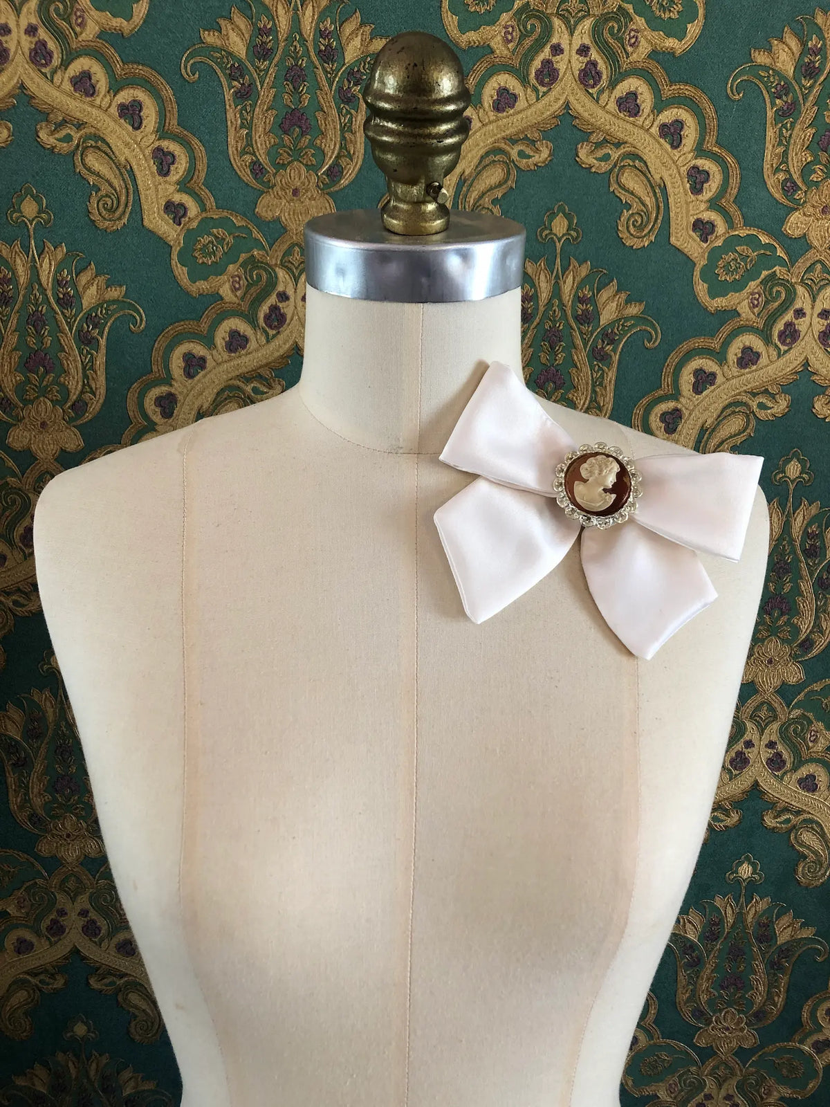 Broochella Camée Ivoire Ivory Cameo Bow Brooch featuring an ivory satin bow with a cream portrait cameo in an ornate silver-tone setting, styled on a cream mannequin against green, gold and purple patterned wallpaper.
