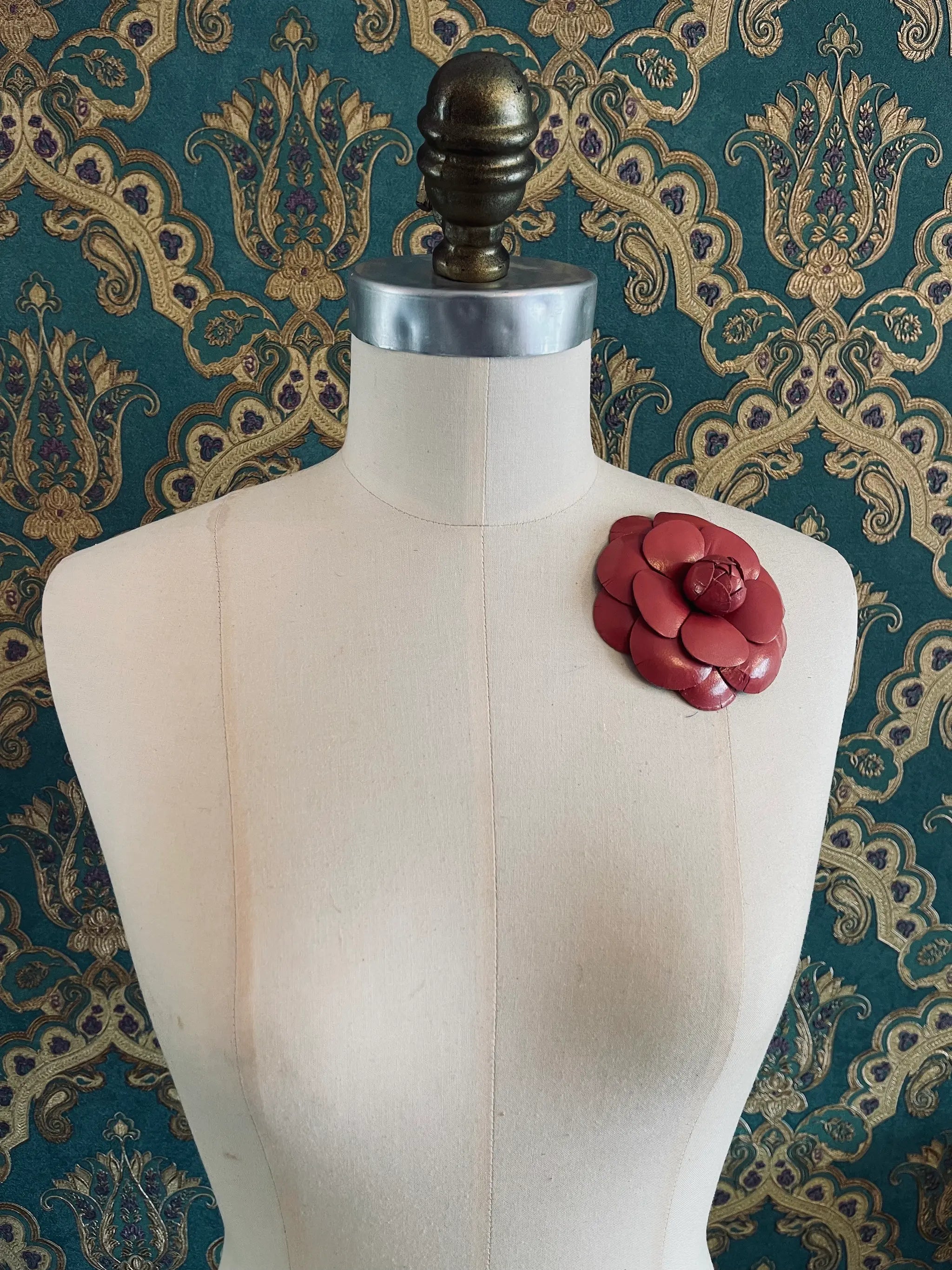 Broochella leather handmade camelia coral designer brooch. Styled on a mannequin. The background is green and gold damask wallpaper