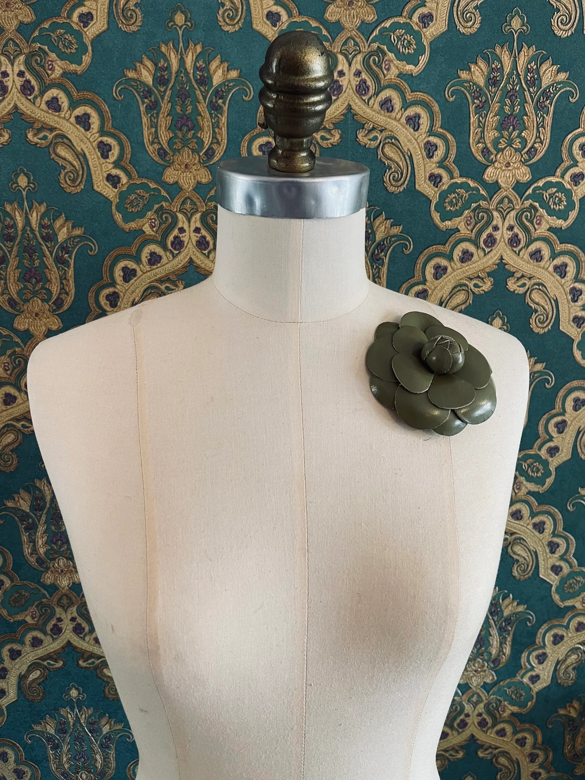 Broochella Camelia handmade, leather, green, designer flower brooch. Styled on a mannequin. The background is green and gold damask wallpaper
