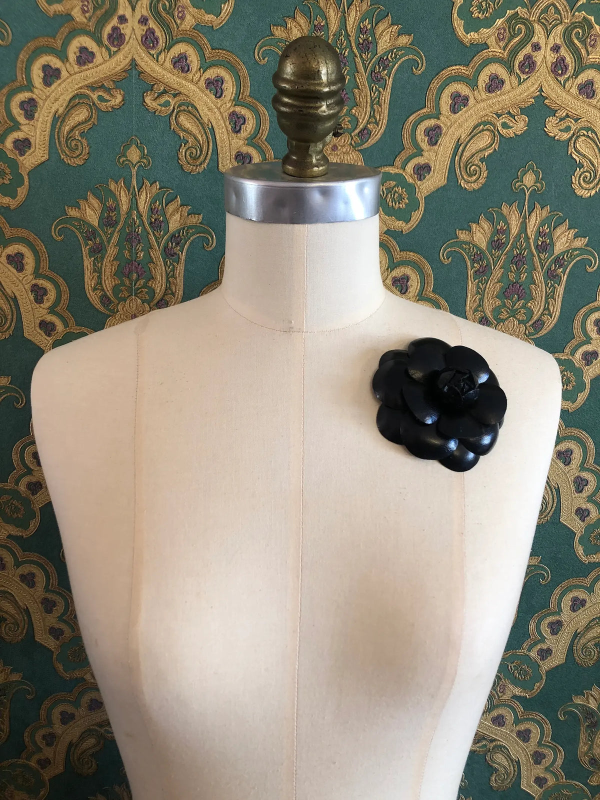 A Broochella Camelia  flower brooch or pin displayed on a cream coloured dress form against a green and gold patterned wallpaper background.
