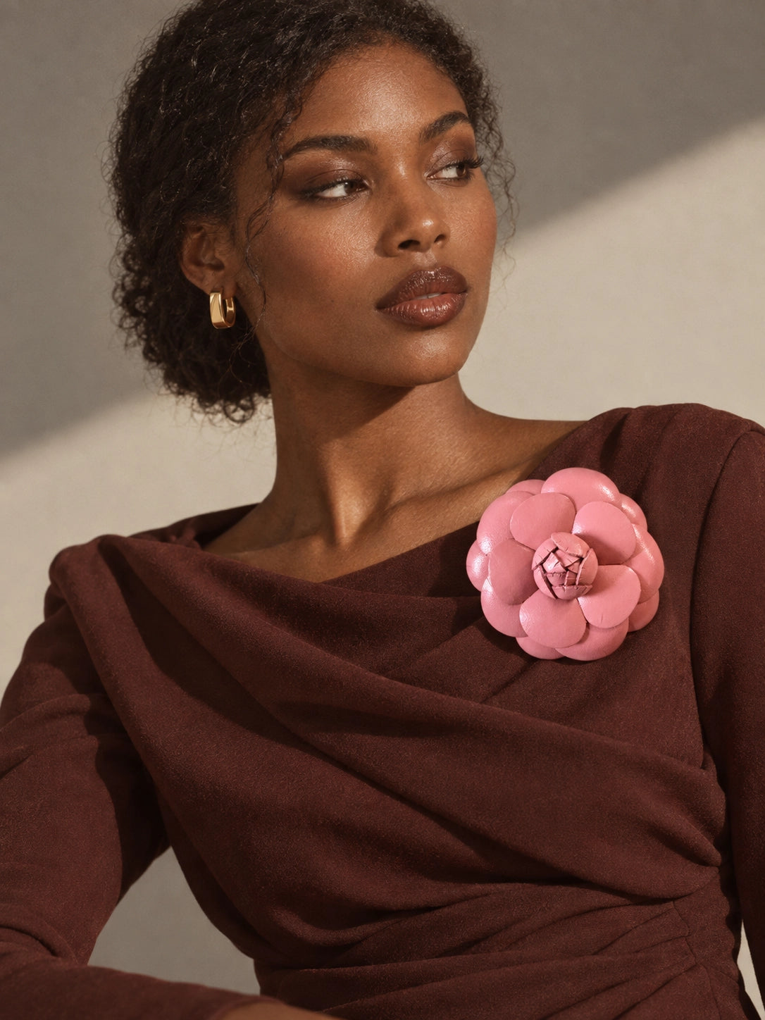 A close-up of a woman wearing a dark brown draped top with a Broochella Camelia leather pink flower-shaped brooch pinned to the chest, set against a warm beige background with dramatic shadows.