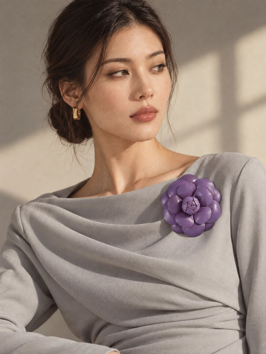 A woman wearing a light gray draped top with a Broochella Camelia leather purple handmade flower-shaped brooch on the shoulder, photographed in warm natural light against a beige wall.
