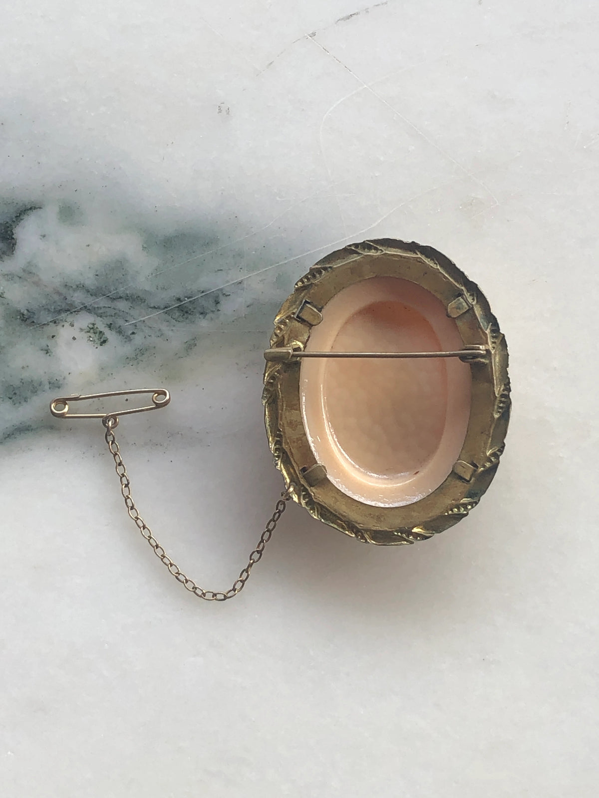 Broochella Cameo della Contessa Vintage Brooch Pin back view showing the antique gold-tone setting, pin fastening and attached safety chain, photographed on white marble with soft grey-green veining.