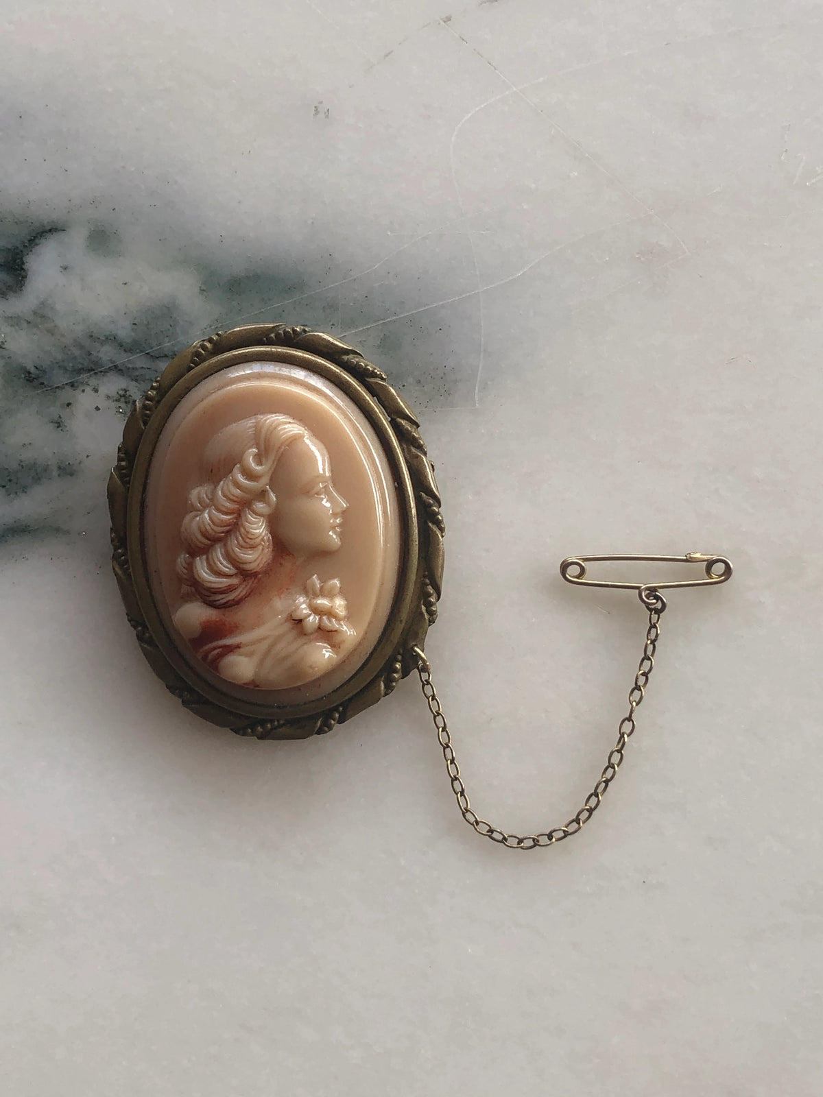 Broochella Cameo della Contessa Vintage Brooch Pin featuring a blush pink carved-style female portrait in an ornate antique gold-tone setting with safety chain and pin, photographed on white marble with soft grey-green veining.
