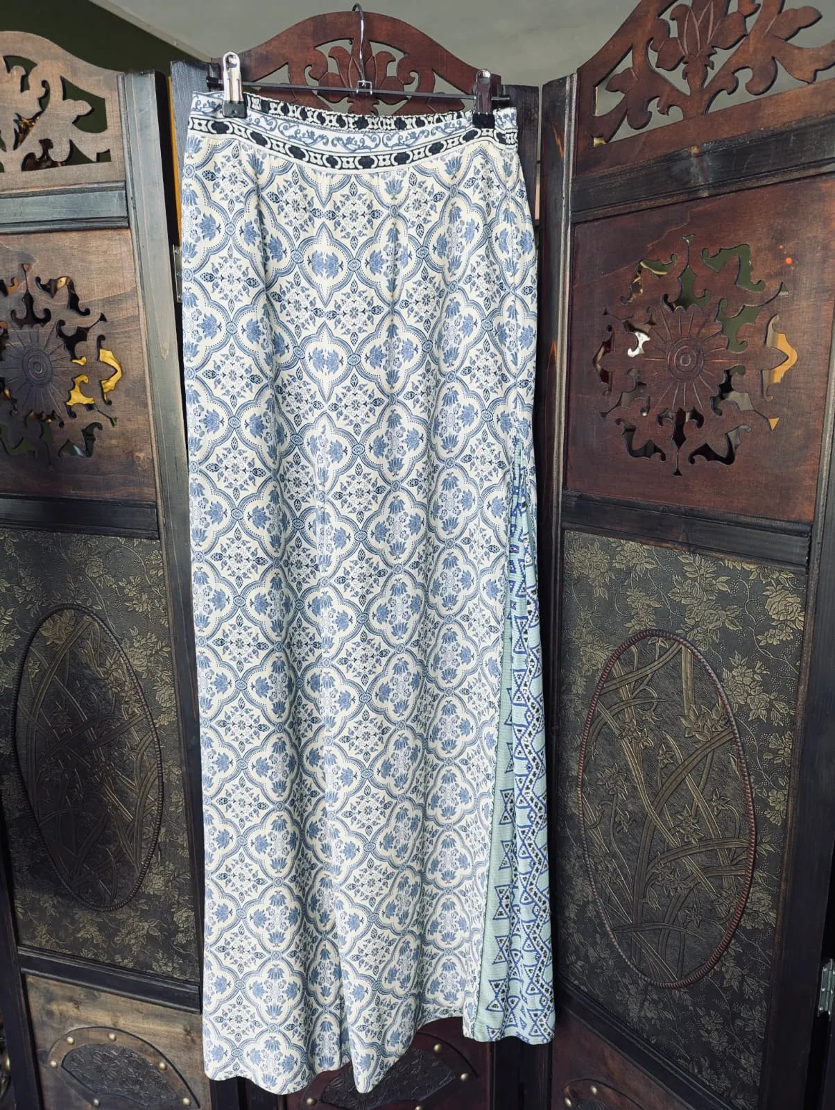 Broochelal Camilla wide leg silk pants in white with a blue tile print. The are pictured hanging on a dark wood room divider. 