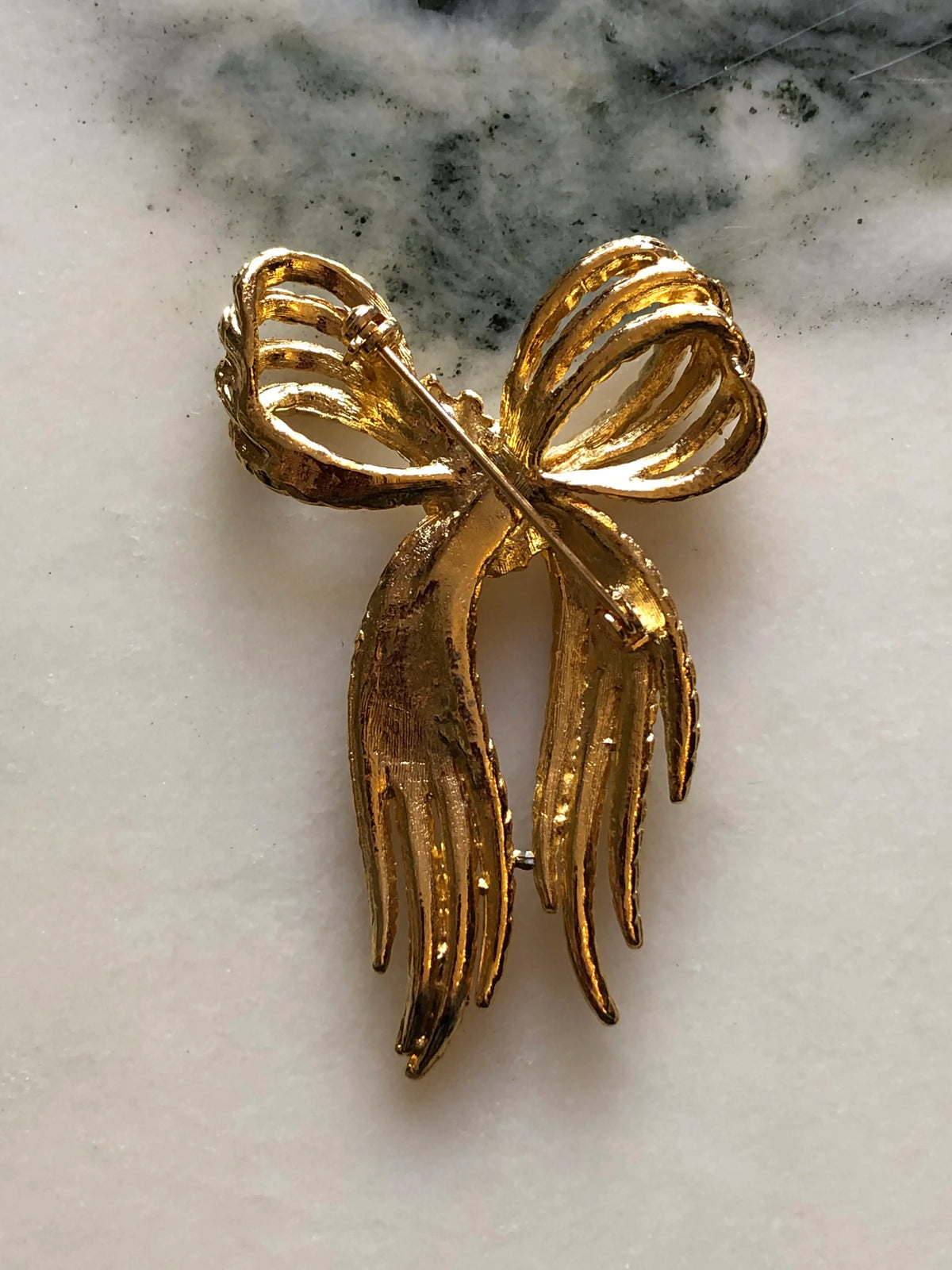 Broochella Camilla Vintage Gold Rope Bow Brooch featuring twisted gold-tone rope loops and long textured ribbon ends, photographed on a cream and grey marble background.