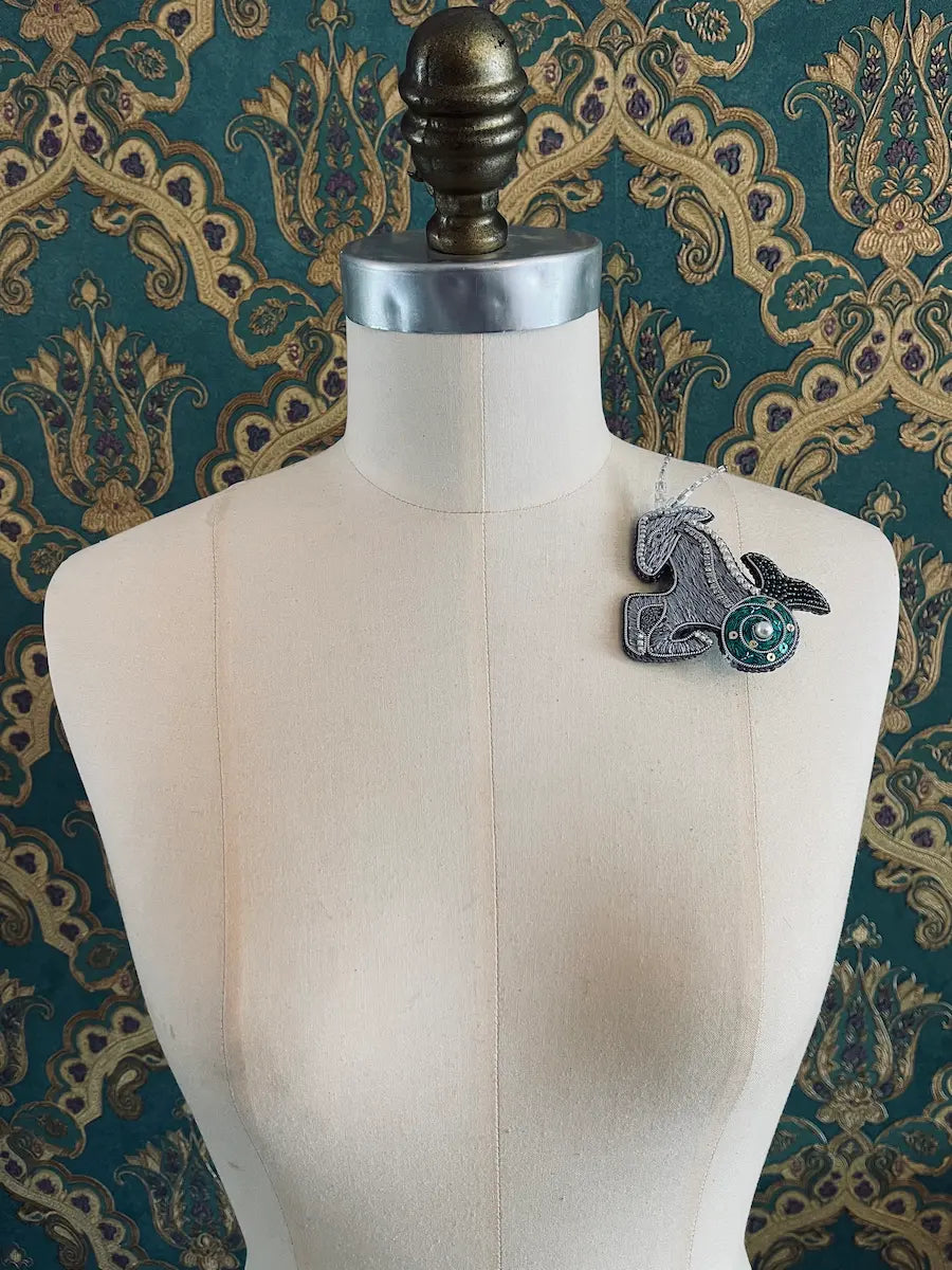 Broochella capricorn hand beaded zodiac designer brooch. Styled on a mannequin. The background is green and gold damask wallpaper