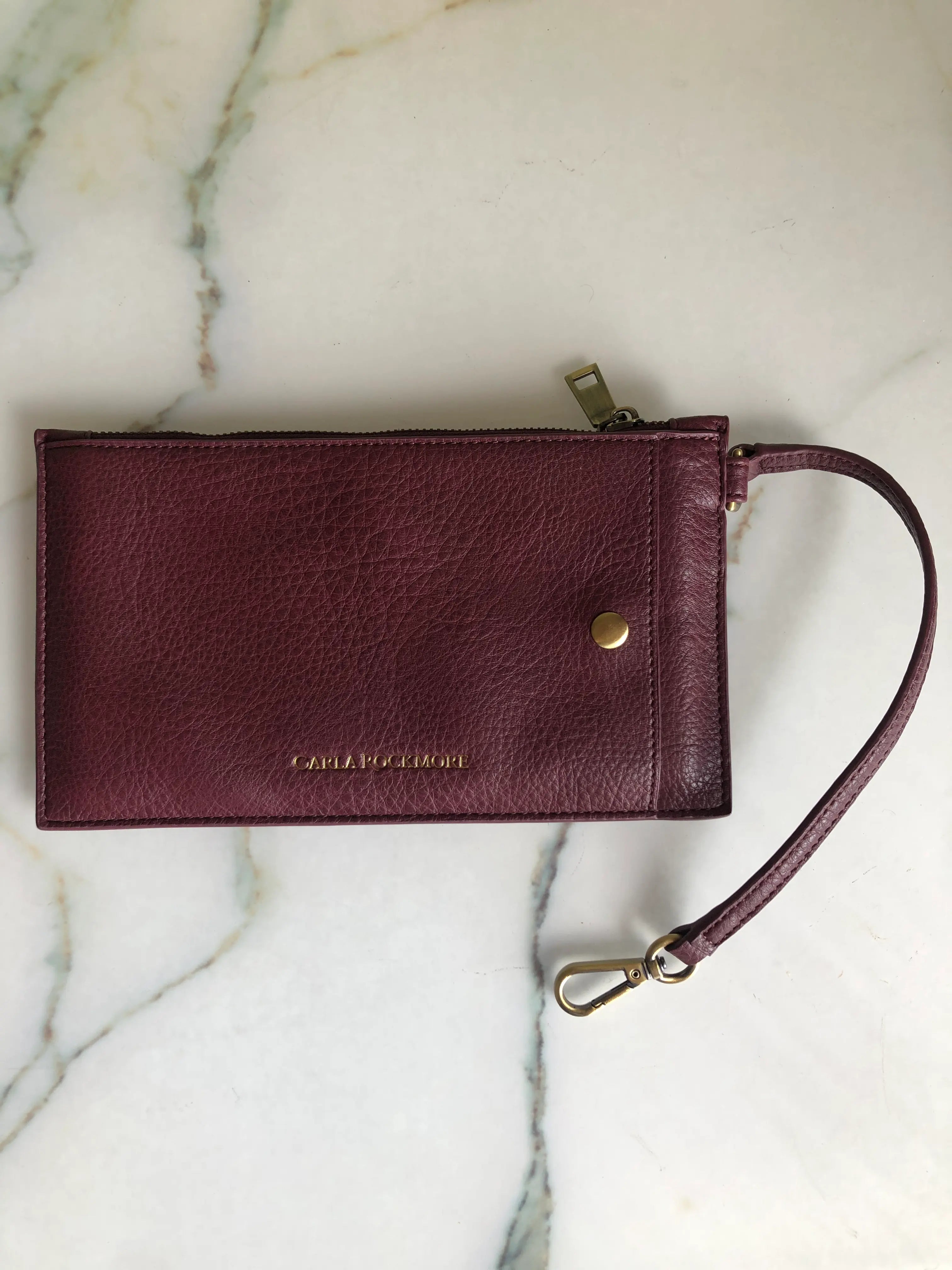 A Broochella Carla Rockmore burgundy pebbled leather wristlet pouch with a gold snap button, gold zipper pull, and a matching wrist strap with a gold clasp, displayed on a white marble surface.