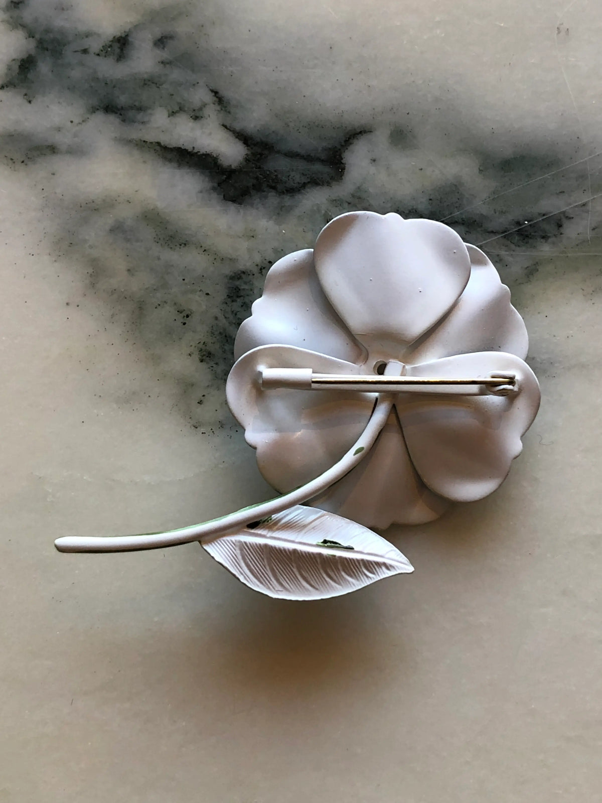 The back view of Broochella Carlotta Vintage Blue Enamel Flower Brooch featuring layered blue enamel petals with black and white detailing, a black centre and sculptural green leaf on a cream and grey marble background.