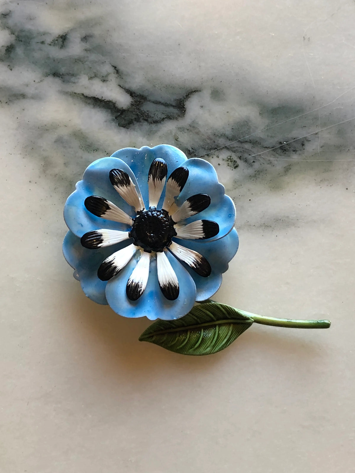 Broochella Carlotta Vintage Blue Enamel Flower Brooch featuring layered blue enamel petals with black and white detailing, a black centre and sculptural green leaf on a cream and grey marble background.