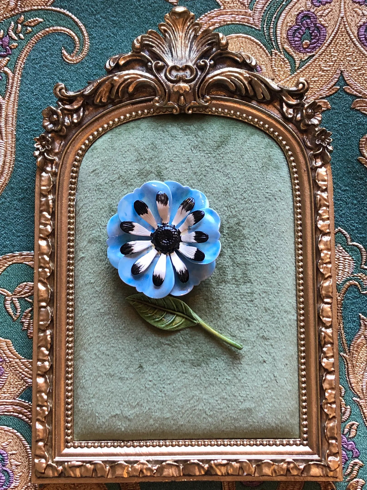 Broochella Carlotta Vintage Blue Enamel Flower Brooch featuring layered blue enamel petals with black and white detailing, a black centre and sculptural green leaf. It is displayed on a green velvet tray with an ornate gold frame. The background is green and gold damask wallpaper