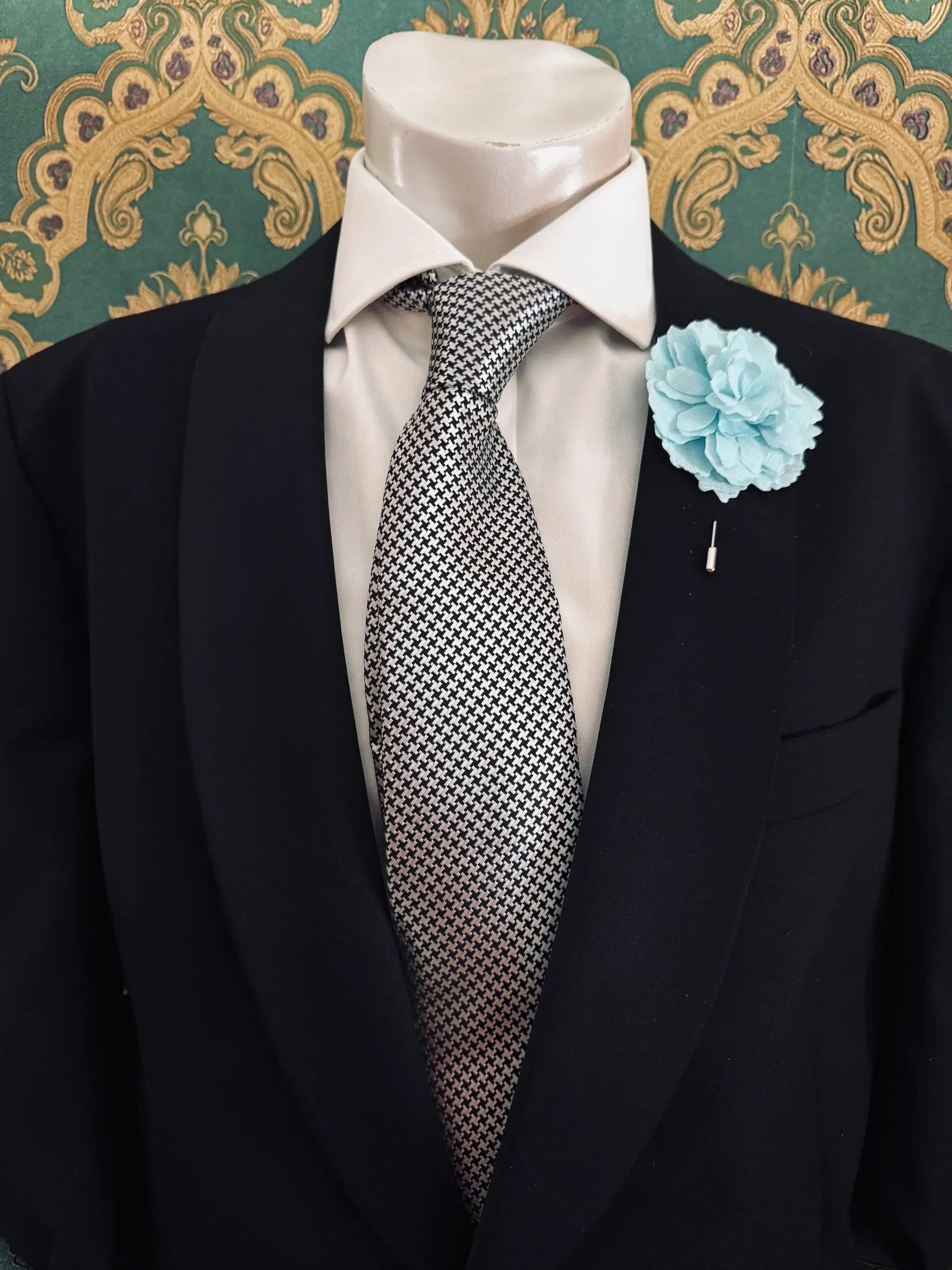 Broochella men's handmade linen carnation brooch in aqua. Pinned to a men's mannequin wearing a black suit and a tie. The background is green and gold damask wallpaper.