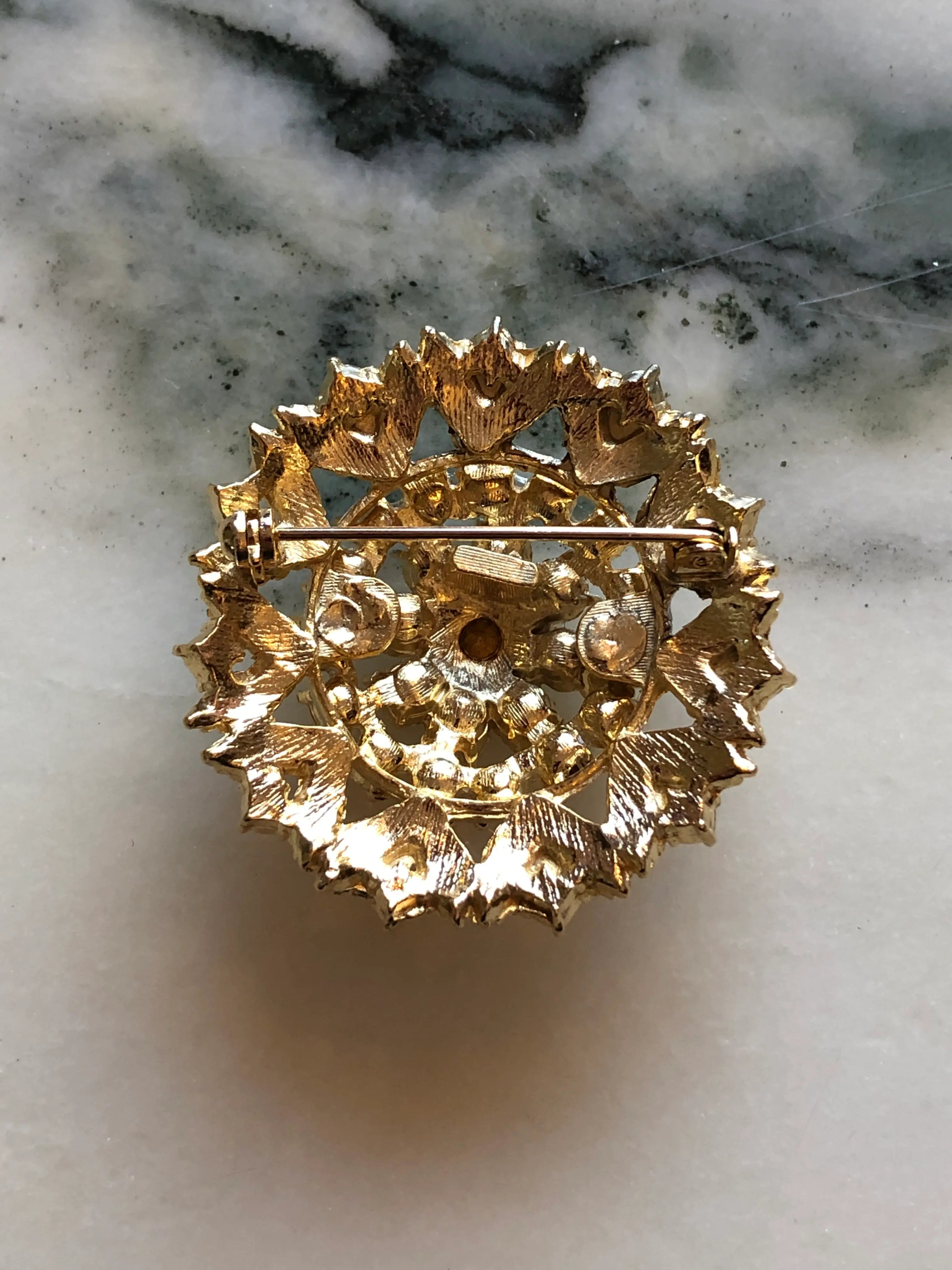 The back view of Broochella Caterina Vintage Gold Crystal Brooch featuring clear faceted crystals in an ornate textured gold-tone circular setting, photographed on a cream and grey marble background.